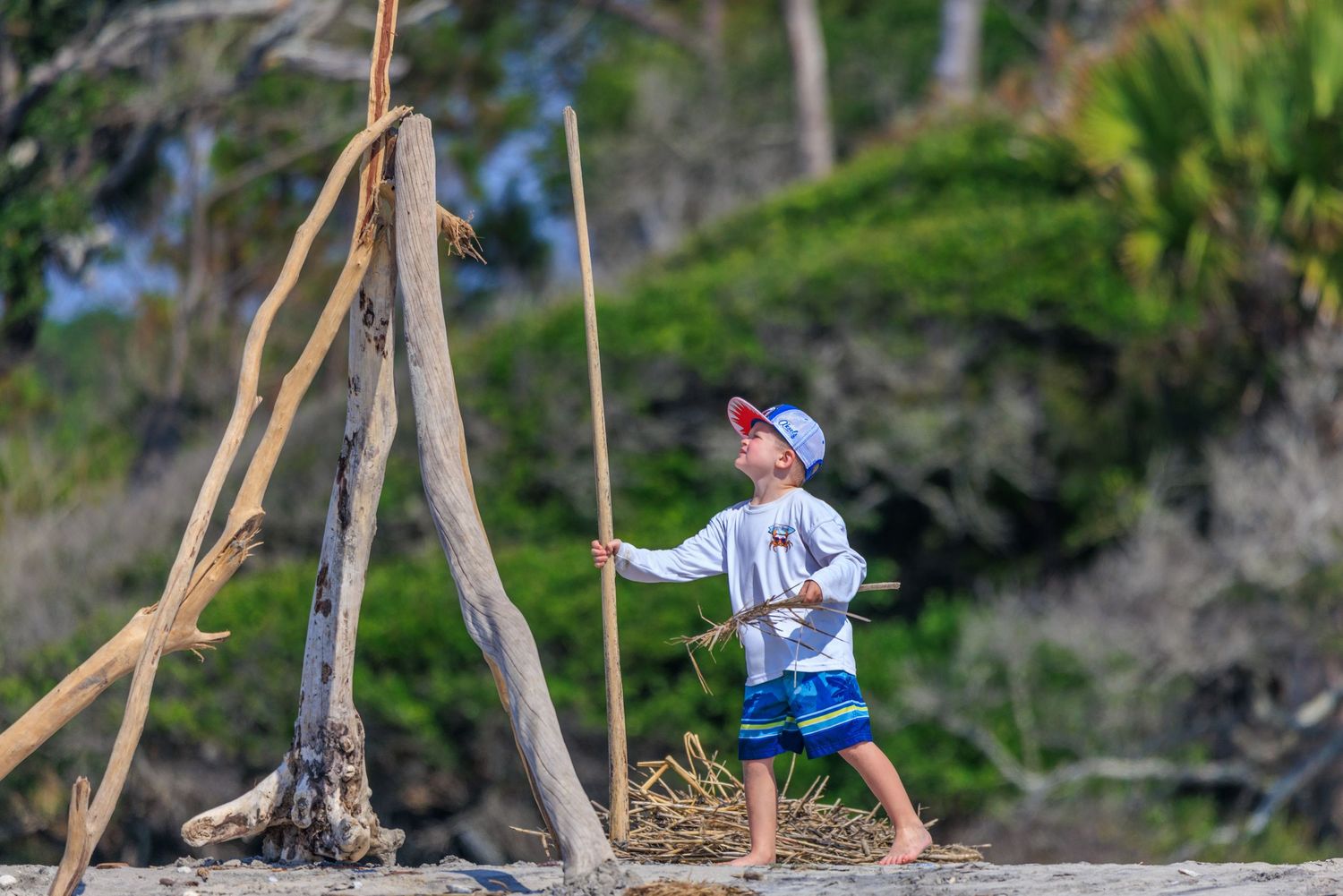 Young boy plays explorer with a wooden stick near driftwood structures on a sandy beach.