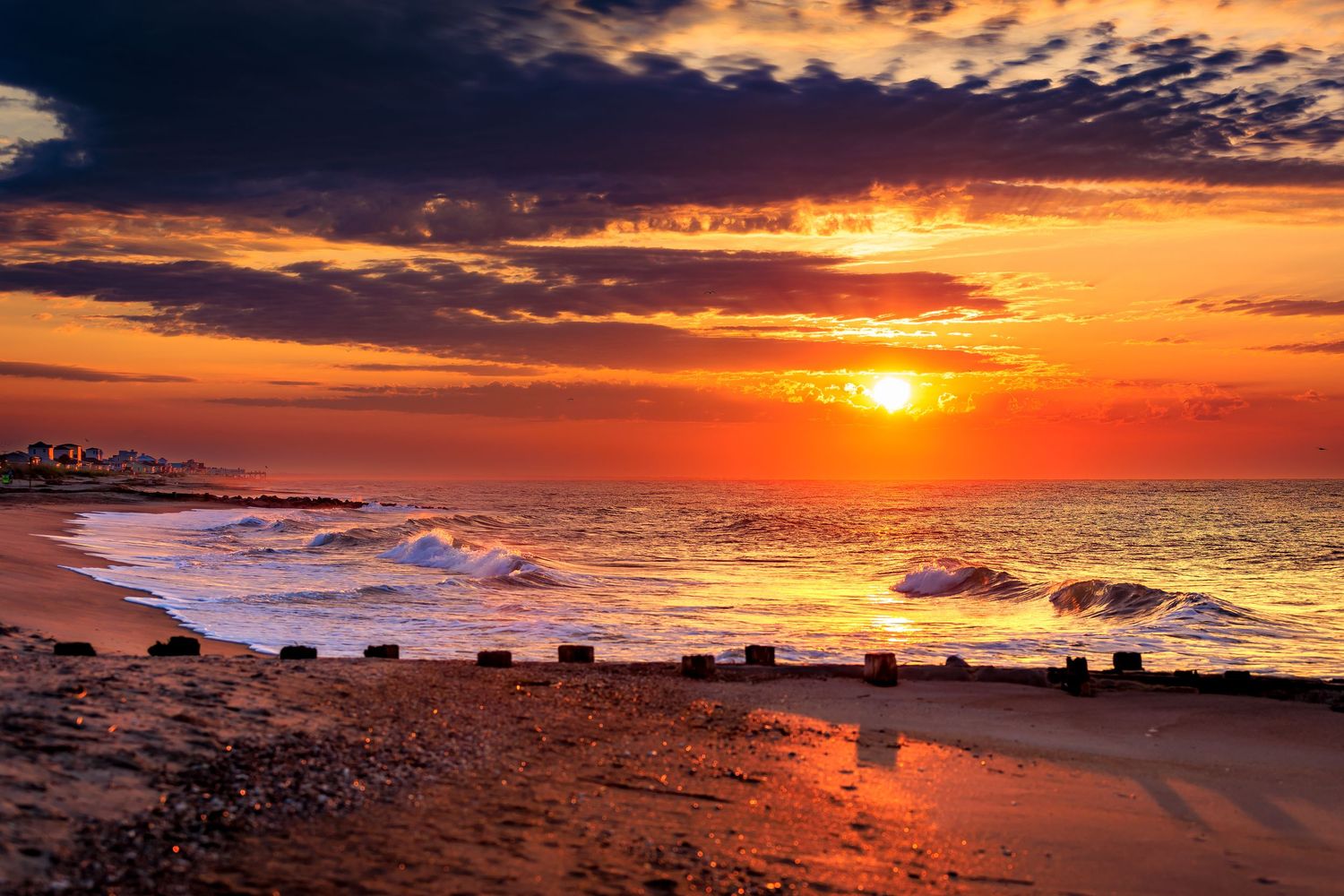 Stunning beach sunset with vivid orange sky, crashing waves, and wooden groyne structures on shore.