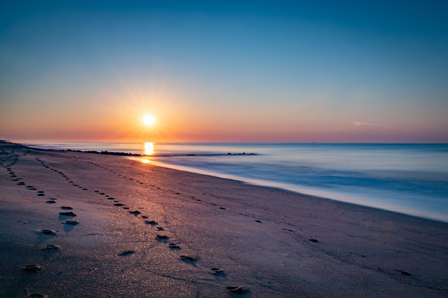 Peaceful sunrise over a sandy beach with footprints along the shore.