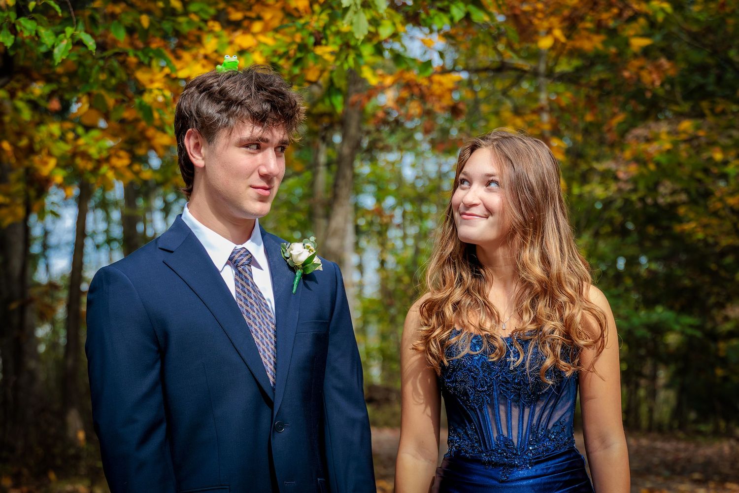 A young couple in formal blue attire poses together outdoors among colorful autumn foliage.