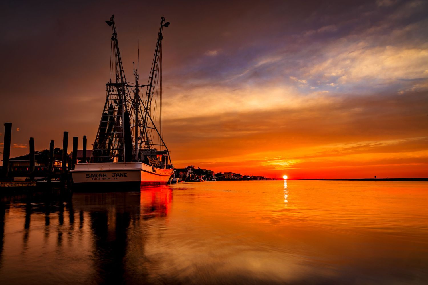 Stunning orange and red sunset reflecting on calm harbor waters with silhouetted shrimp boat docked at pier.