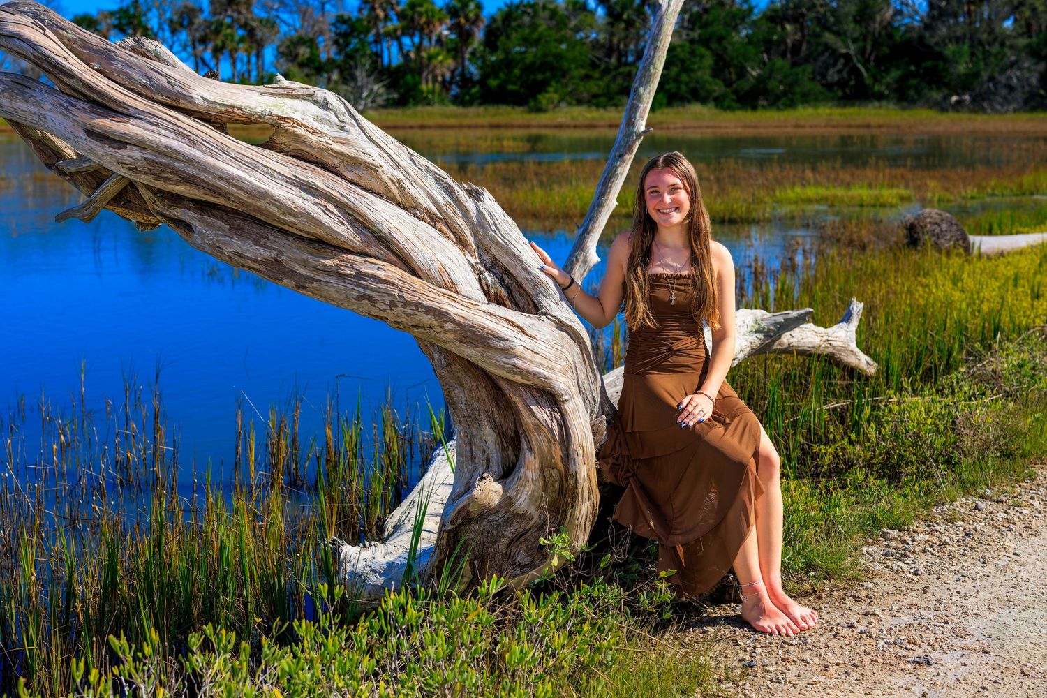 Young woman in brown dress sitting on driftwood by a serene blue lake surrounded by lush greenery.