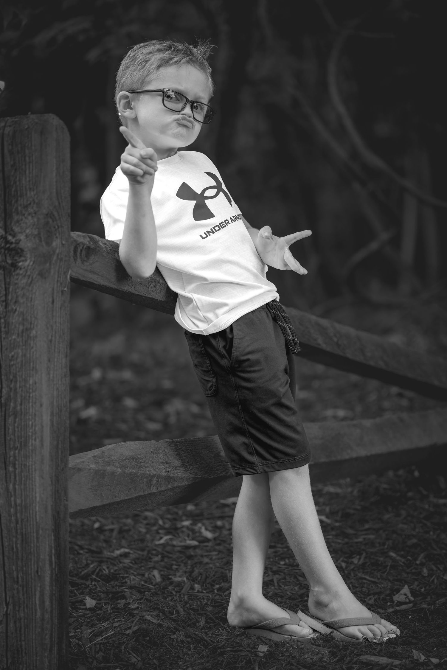 Black and white photo of a young boy in glasses and Under Armour shirt posing confidently by a wooden fence.