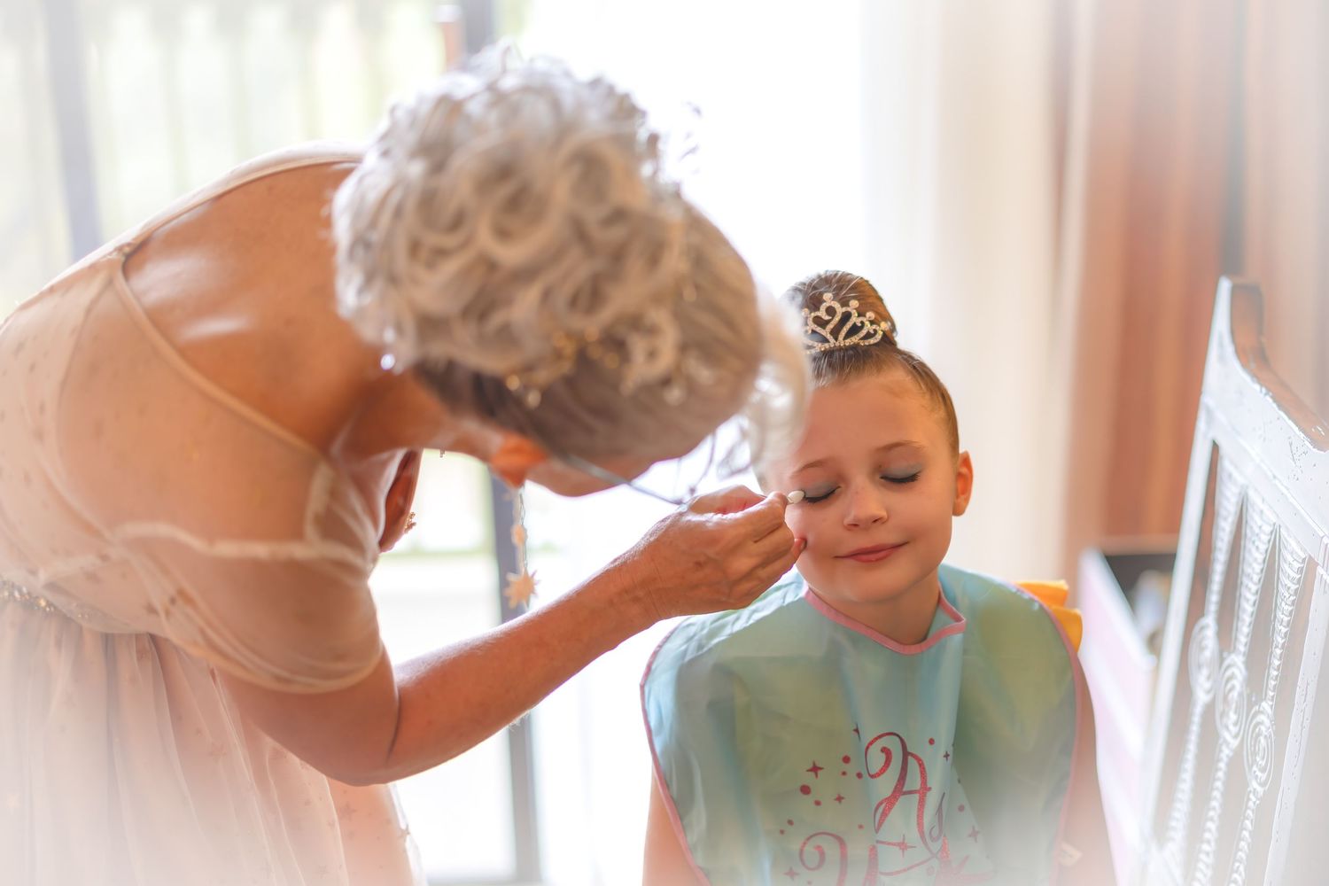 Ballet dancer applies makeup to a young girl wearing a tiara and bib before a dance performance.
