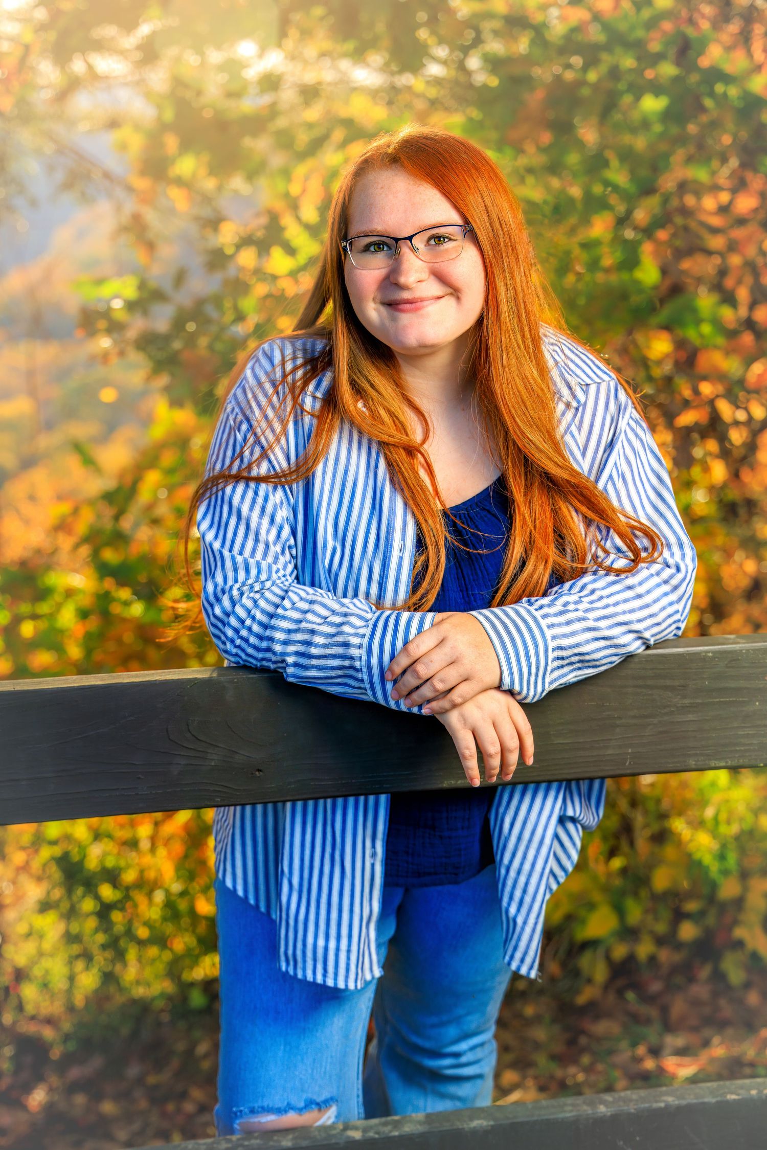 Young redhead woman with glasses smiling outdoors leaning on wooden fence with colorful autumn foliage background.