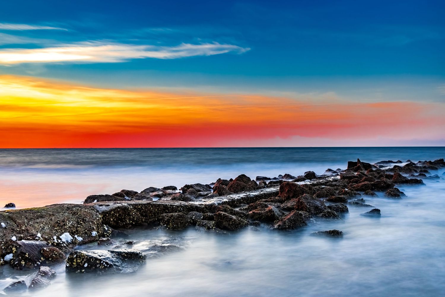 A vibrant sunset over rocky coastline with colorful sky reflecting on calm ocean waters.