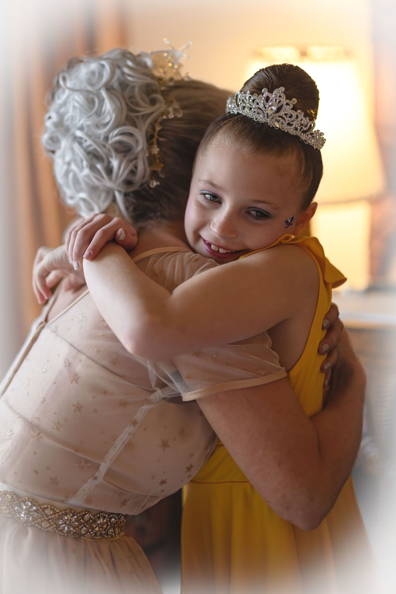 A young girl wearing a tiara and yellow dress shares a warm hug with a woman in a beige gown.