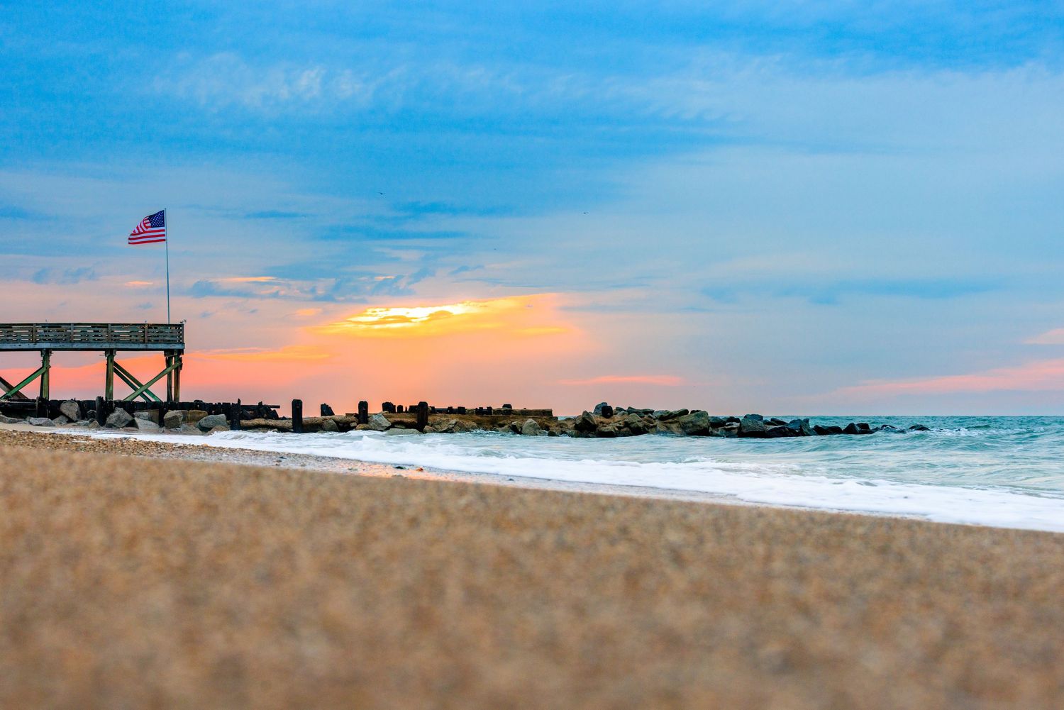 Sandy beach at sunset with an American flag on a pier and rocky jetty nearby.
