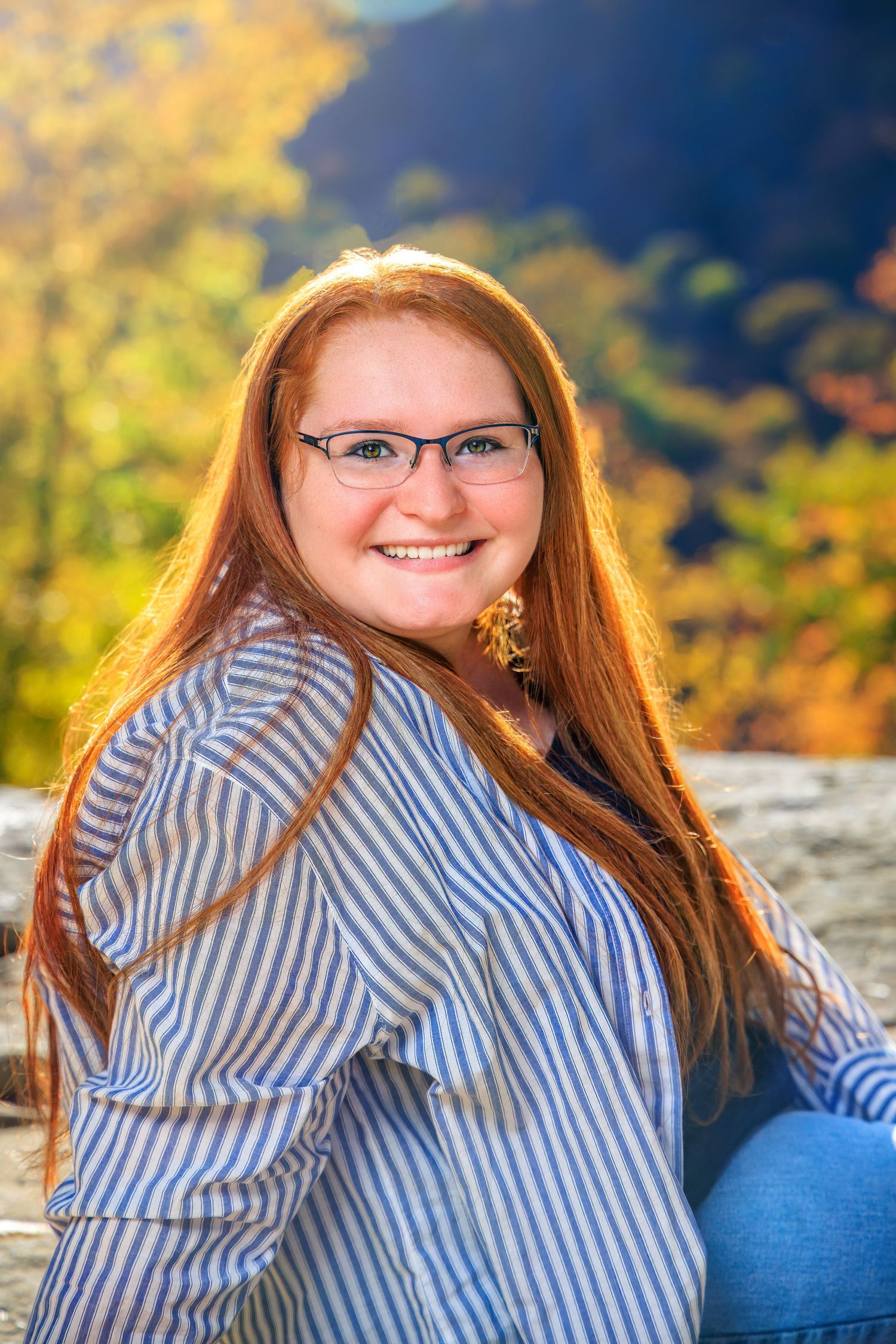Smiling redhead teen girl with glasses poses outdoors against vibrant autumn foliage background.
