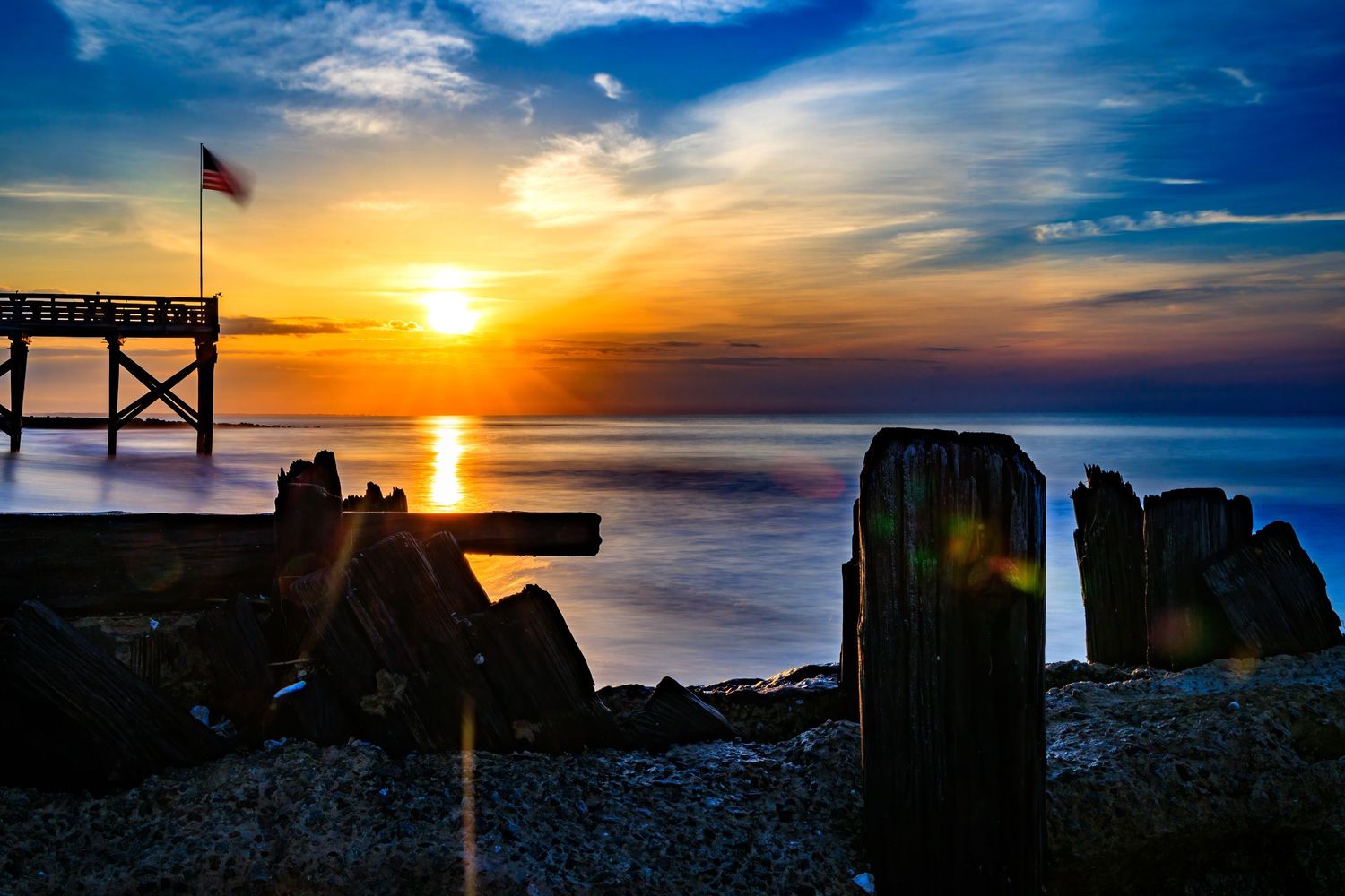 Golden sunset over rocky shoreline with pier and American flag.