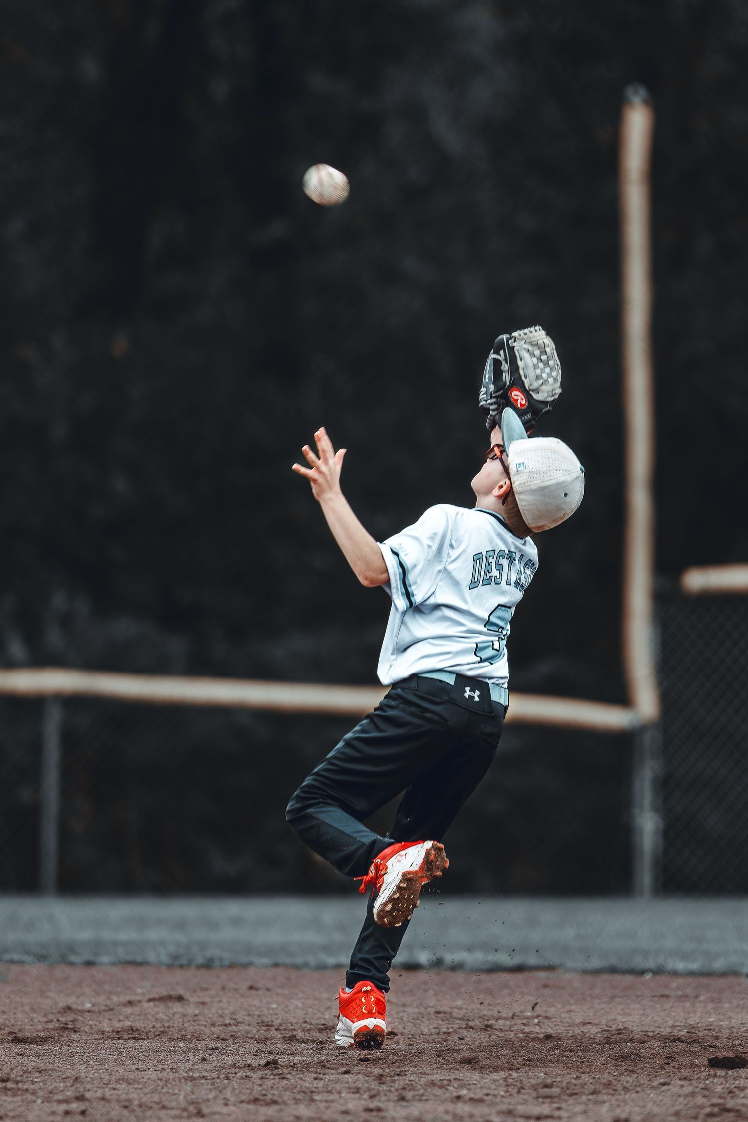 Young baseball player in white uniform reaching up with glove to catch a fly ball on the field.