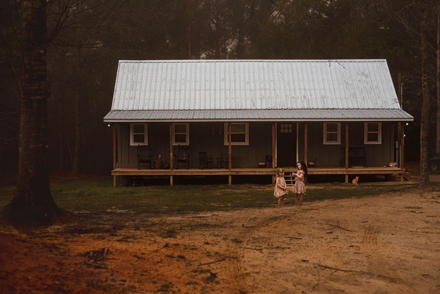 Two girls in dresses stand before their house in the distance during a documentary-style family photography session in Hurley, MS
