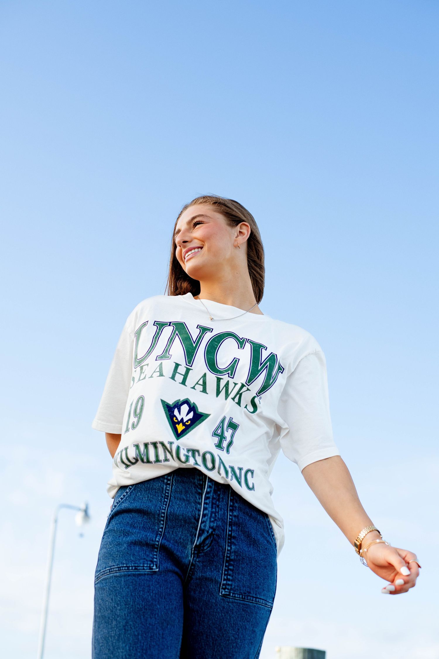 Young woman smiling in white graphic tee and blue jeans against a clear blue sky. Senior session in Gulfport, Mississippi. Unique