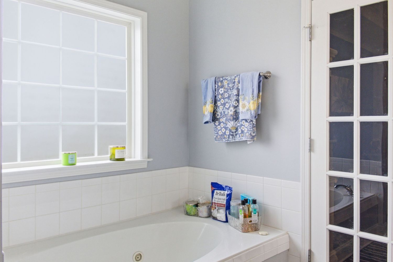 White tiled bathroom with soaking tub, frosted window, and blue towels on wall rack.