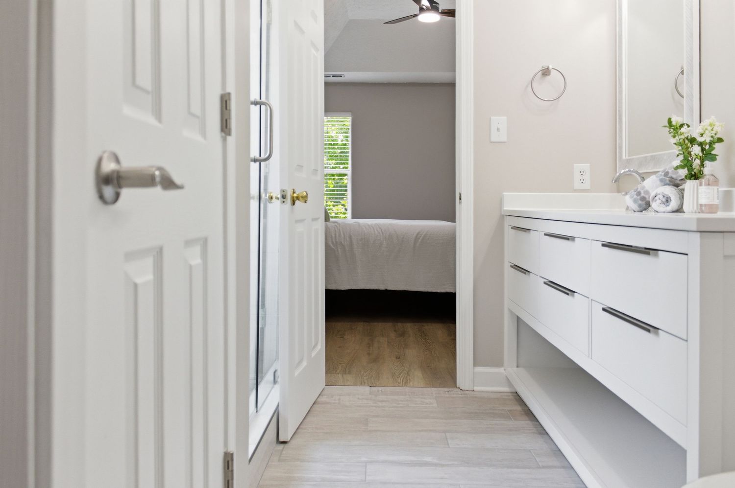Bright white bathroom with floating vanity, marble floors, open door leading to bedroom with ceiling fan.