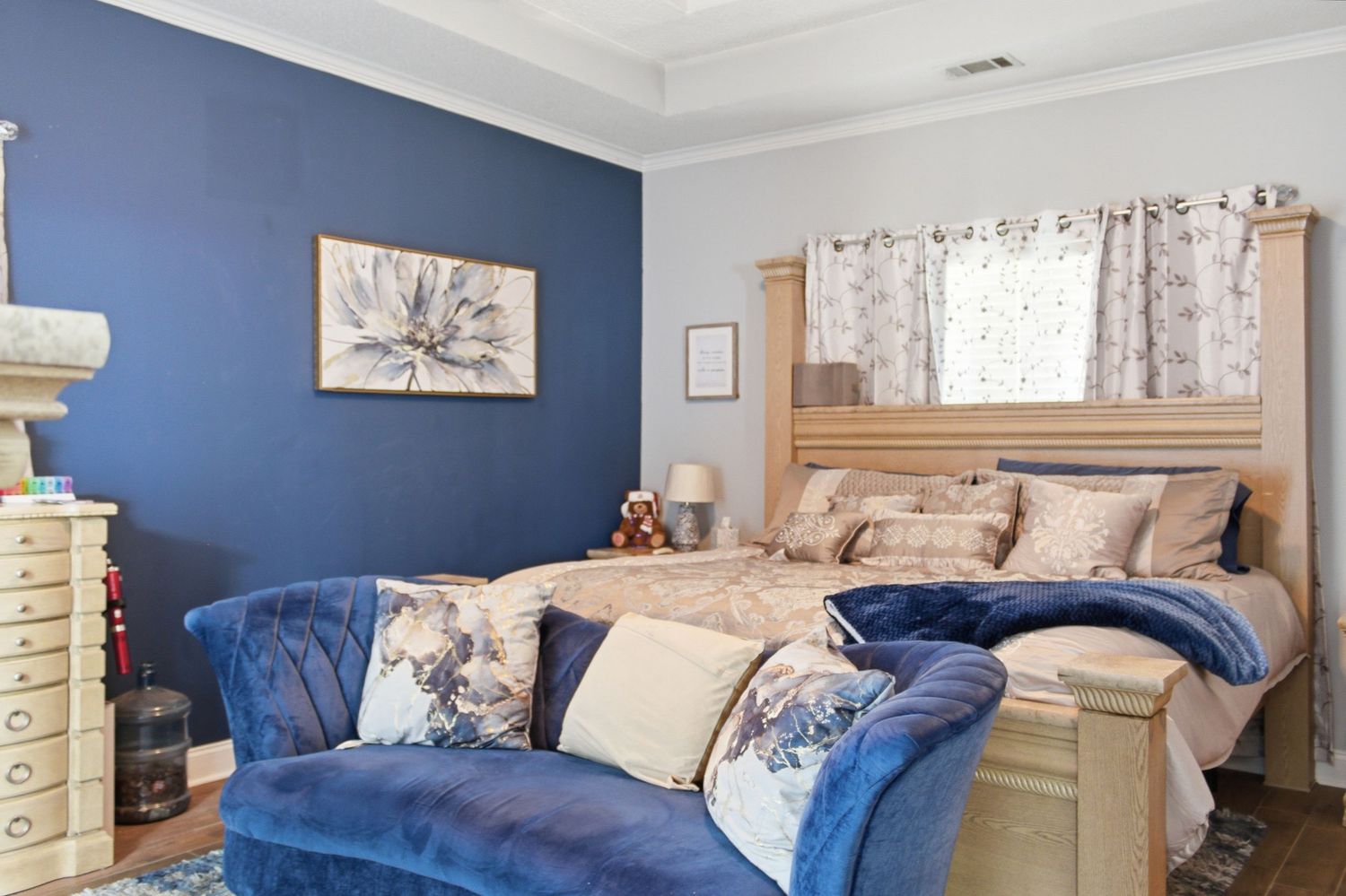 Master bedroom with navy accent wall, wooden bed frame, and blue velvet chaise lounge.