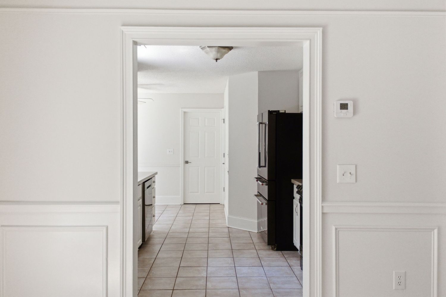 Bright hallway with white wainscoting leading to a kitchen with black refrigerator and white door.