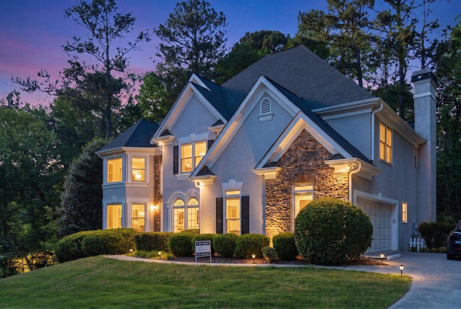 Elegant two-story home with stone facade illuminated at dusk with warm interior and exterior lighting.