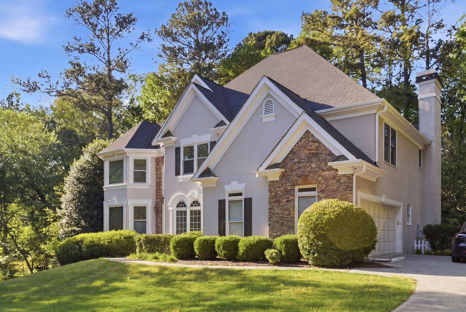 Elegant two-story home with stone and siding exterior, manicured shrubs, and lush green lawn surrounded by tall trees.