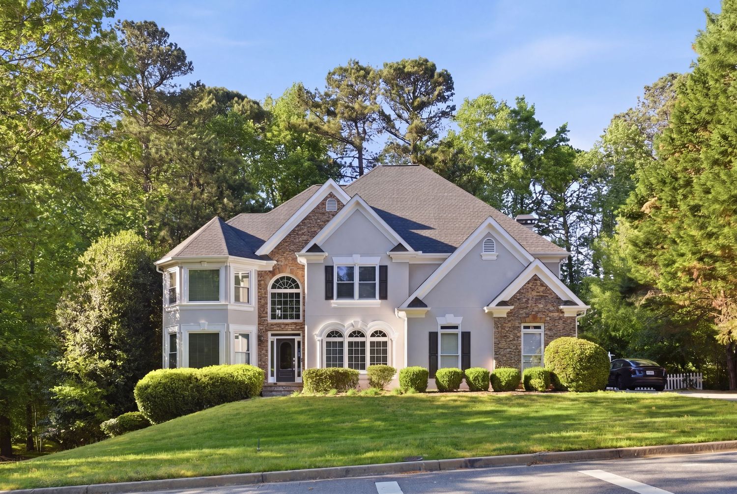 Large two-story brick and stucco home with arched windows, manicured landscaping, and tall trees in a suburban neighborhood.