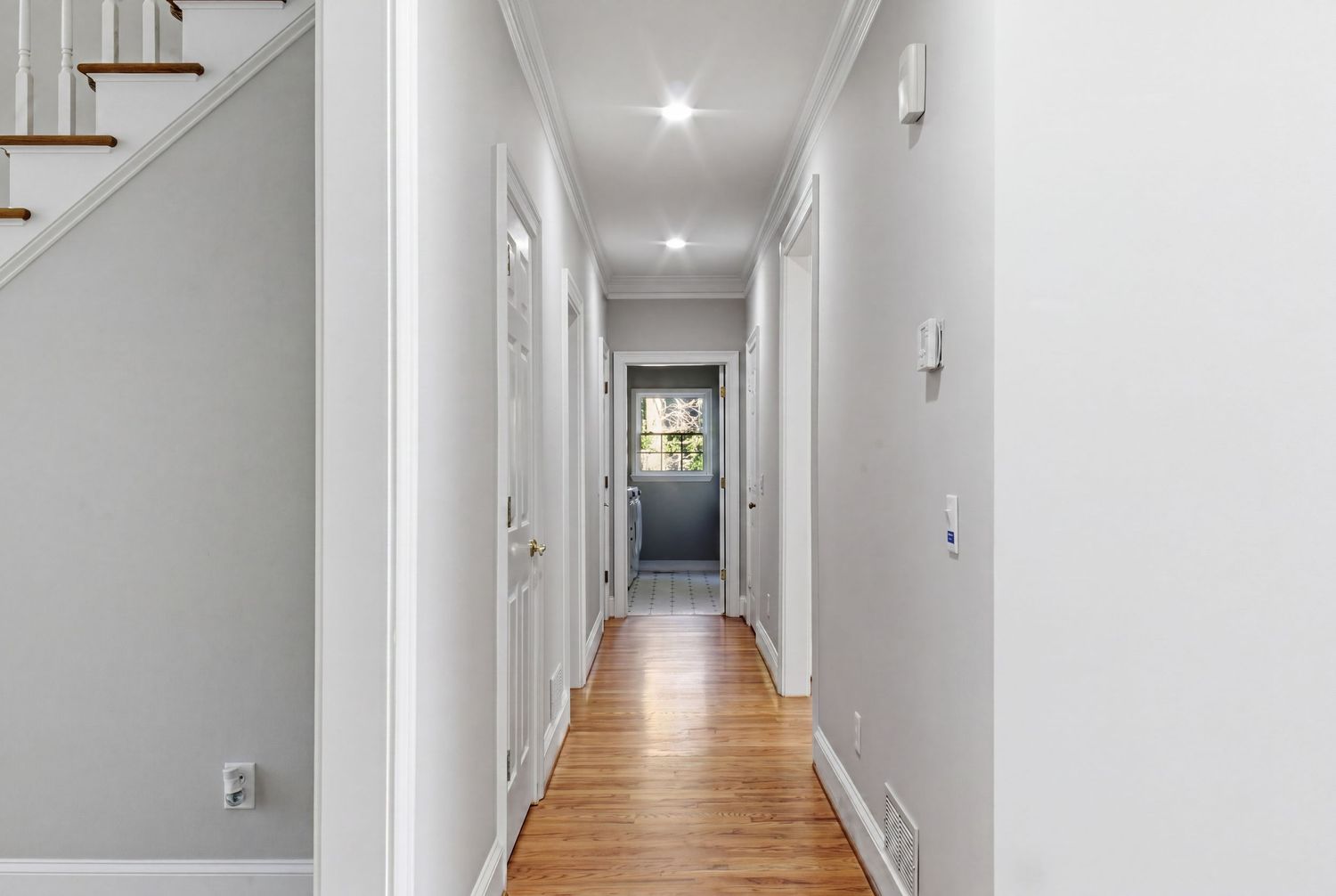 Bright narrow hallway with white walls, hardwood floors, recessed lighting, and white doors leading to a front entrance.