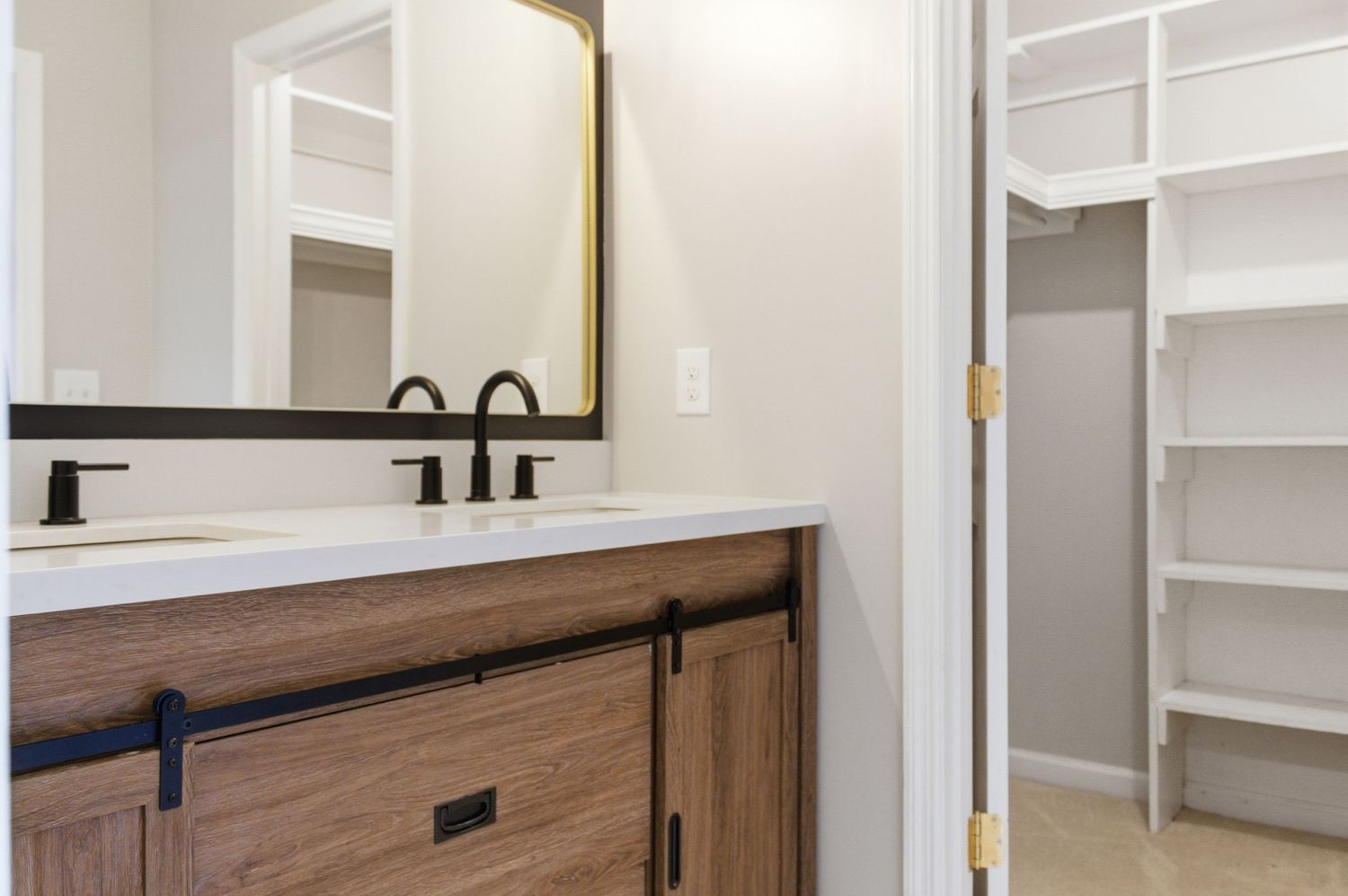 Bathroom vanity with wood cabinet, white countertop, black fixtures, gold mirror, and walk-in closet beyond.