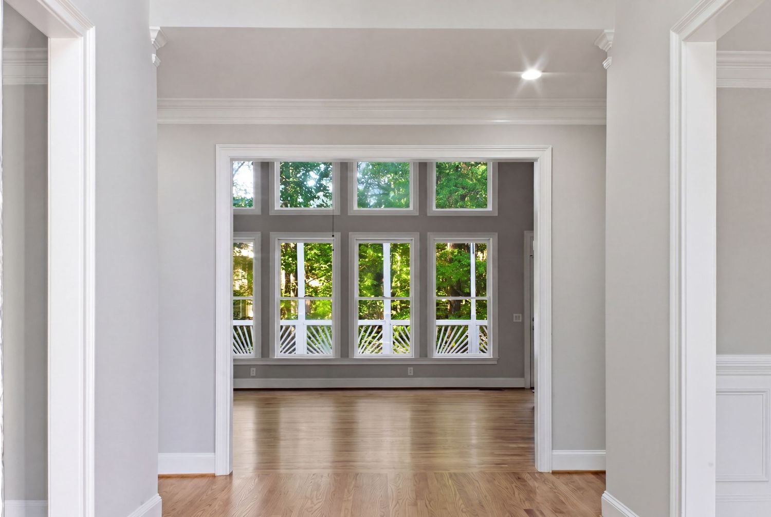 Bright hallway leading to a sunlit room with large French doors, transom windows, and hardwood floors in a modern home.