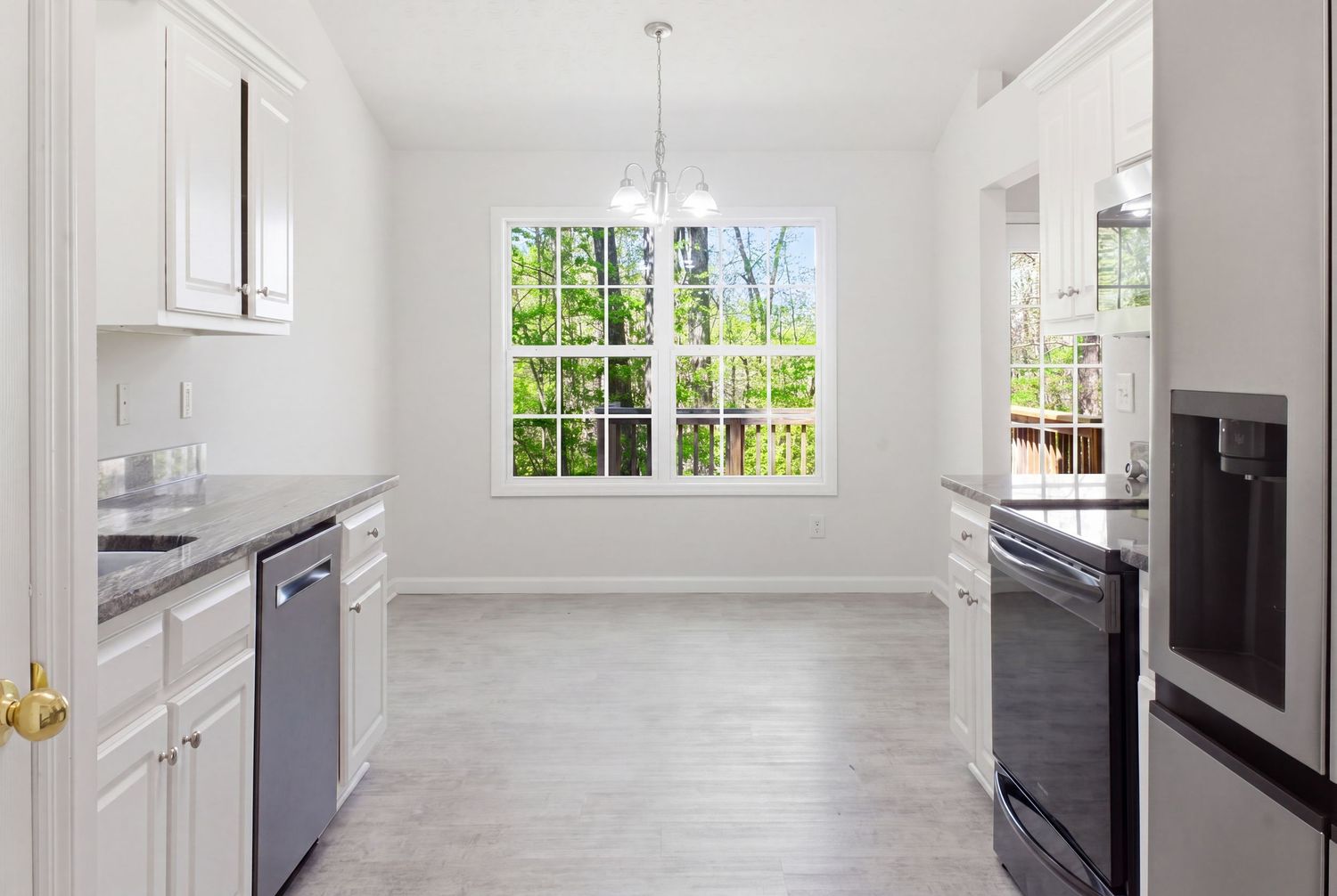 Bright white kitchen with stainless appliances, gray countertops, and a window overlooking lush greenery.