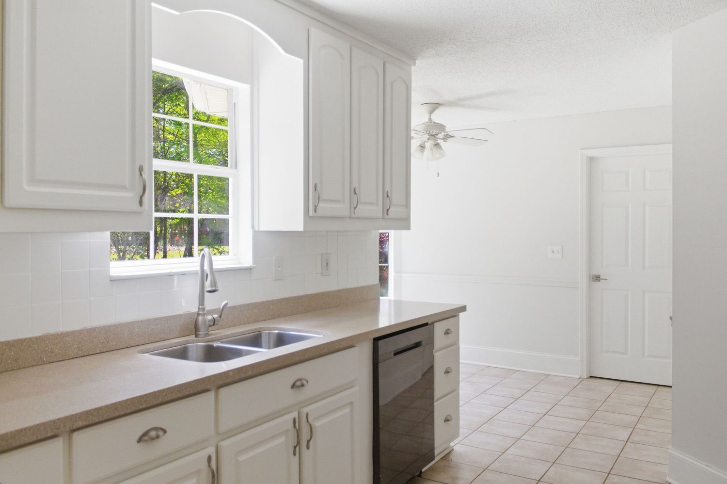 White kitchen with double sink, tan countertops, white cabinets, dishwasher, ceiling fan, and tile flooring.
