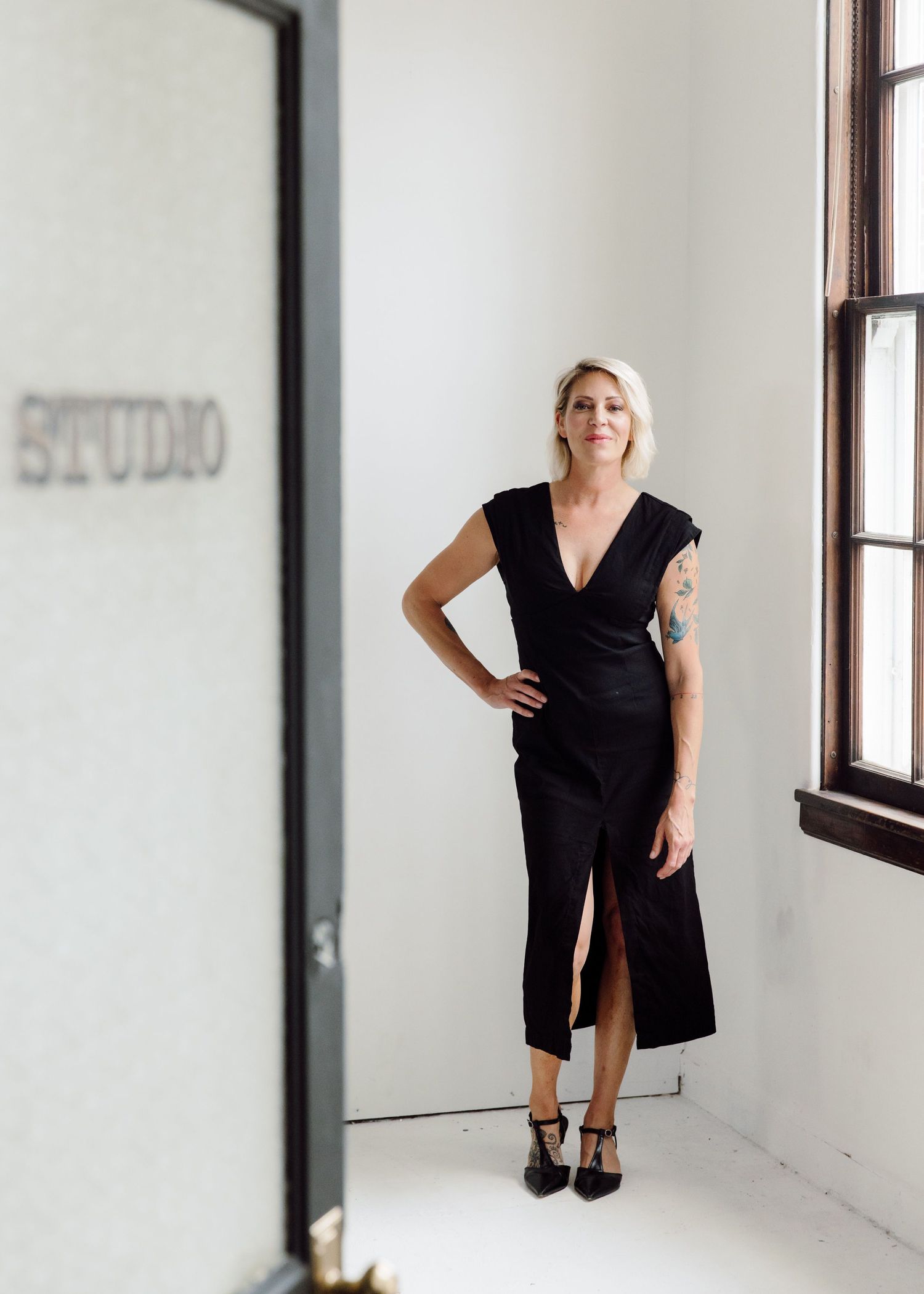 Woman in black wrap dress with tattoo poses confidently in bright studio space with large windows.