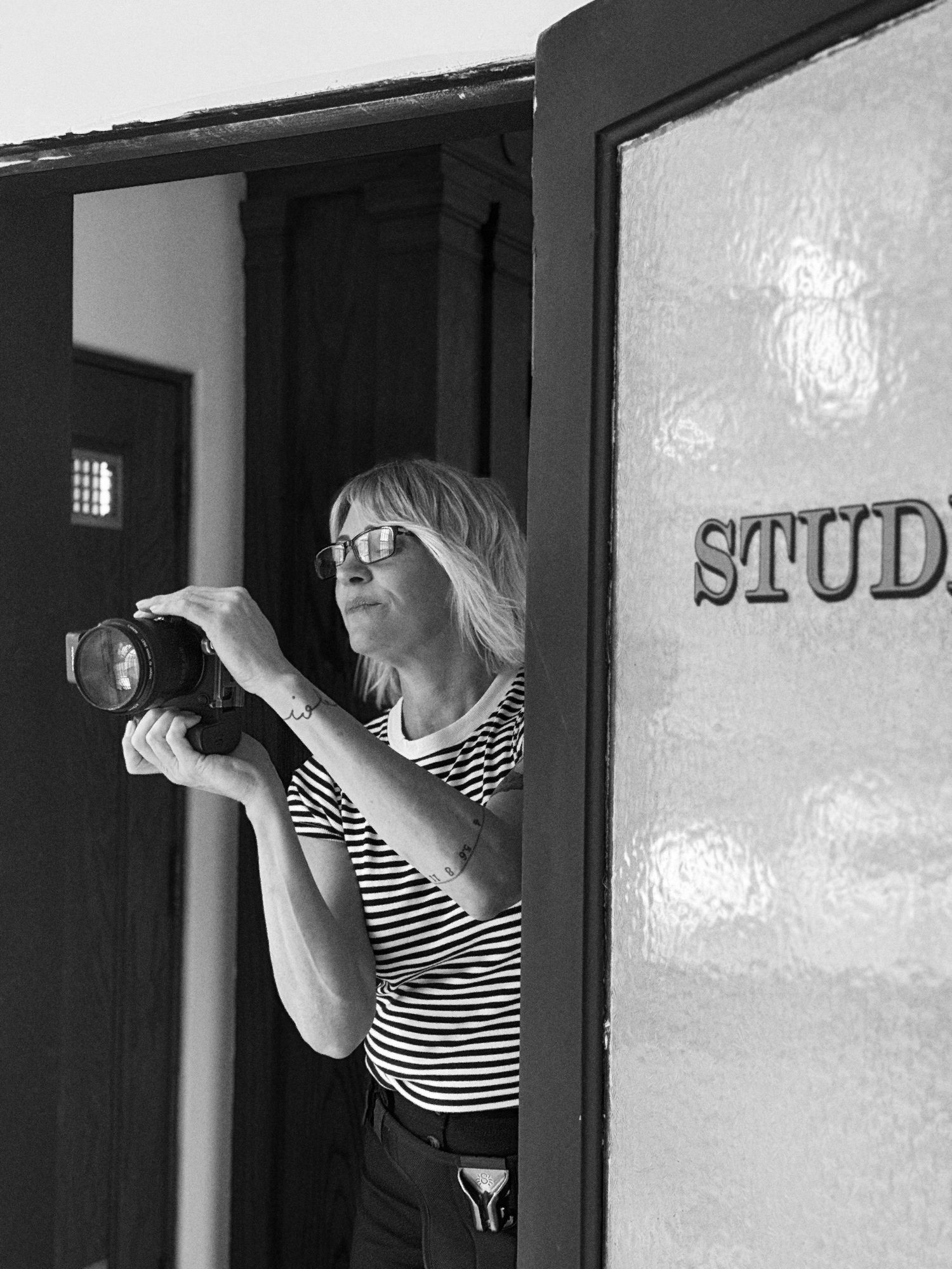 Woman with glasses holding a camera near a studio door, captured in black and white photography.