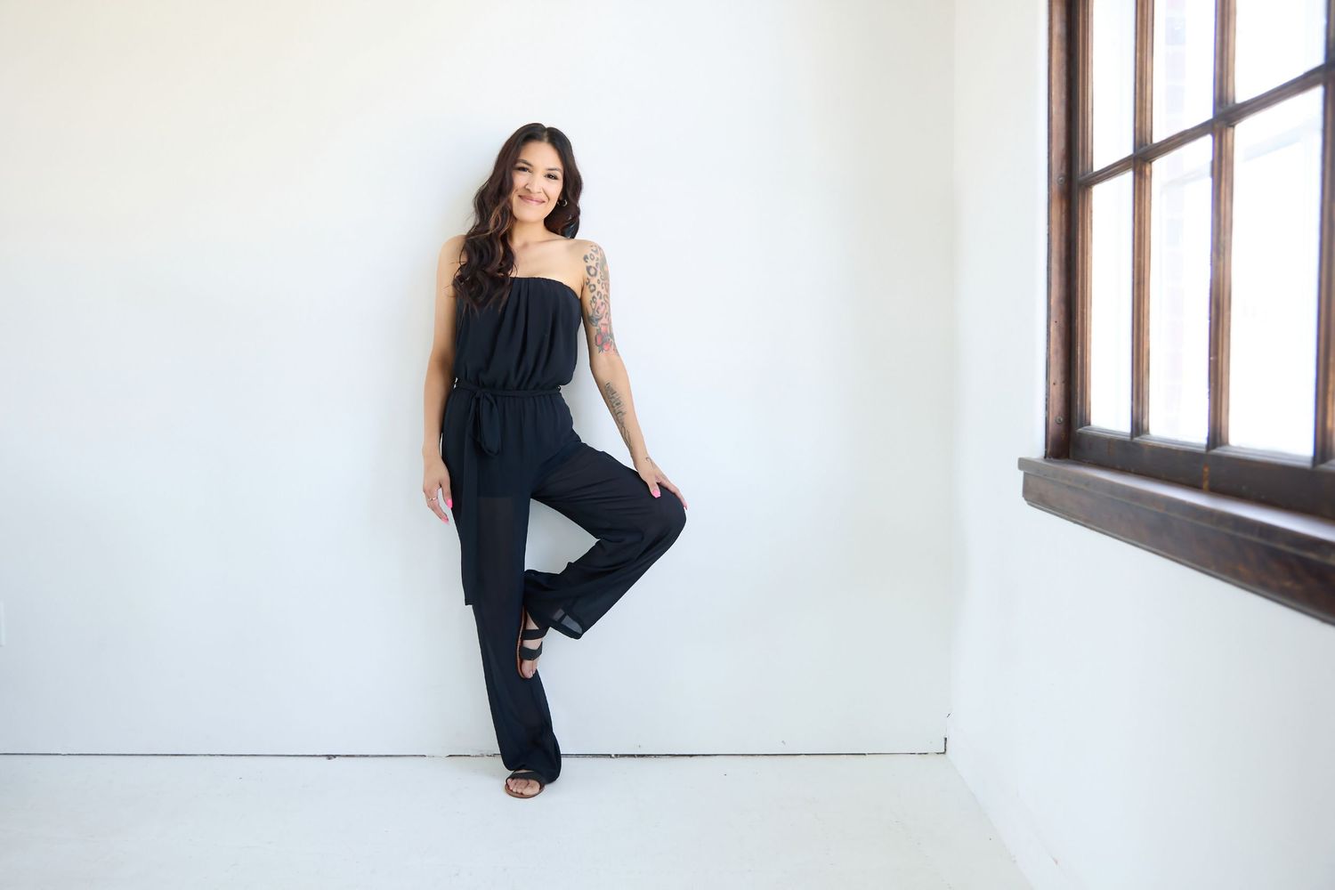 Woman in black strapless jumpsuit posing confidently against a white wall near a window.