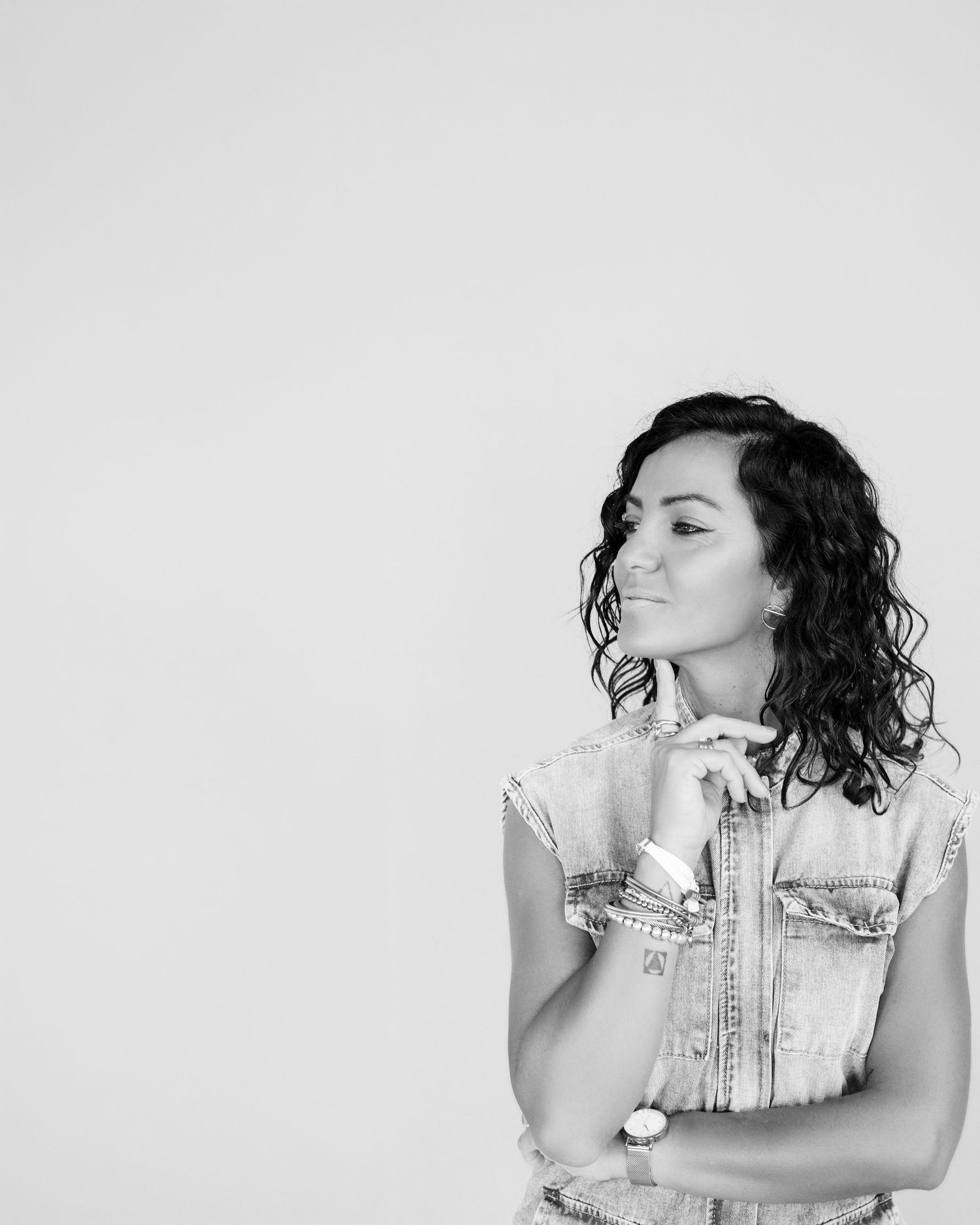 Black and white photo of a young woman with curly hair in a denim vest, posing thoughtfully with hand on chin.