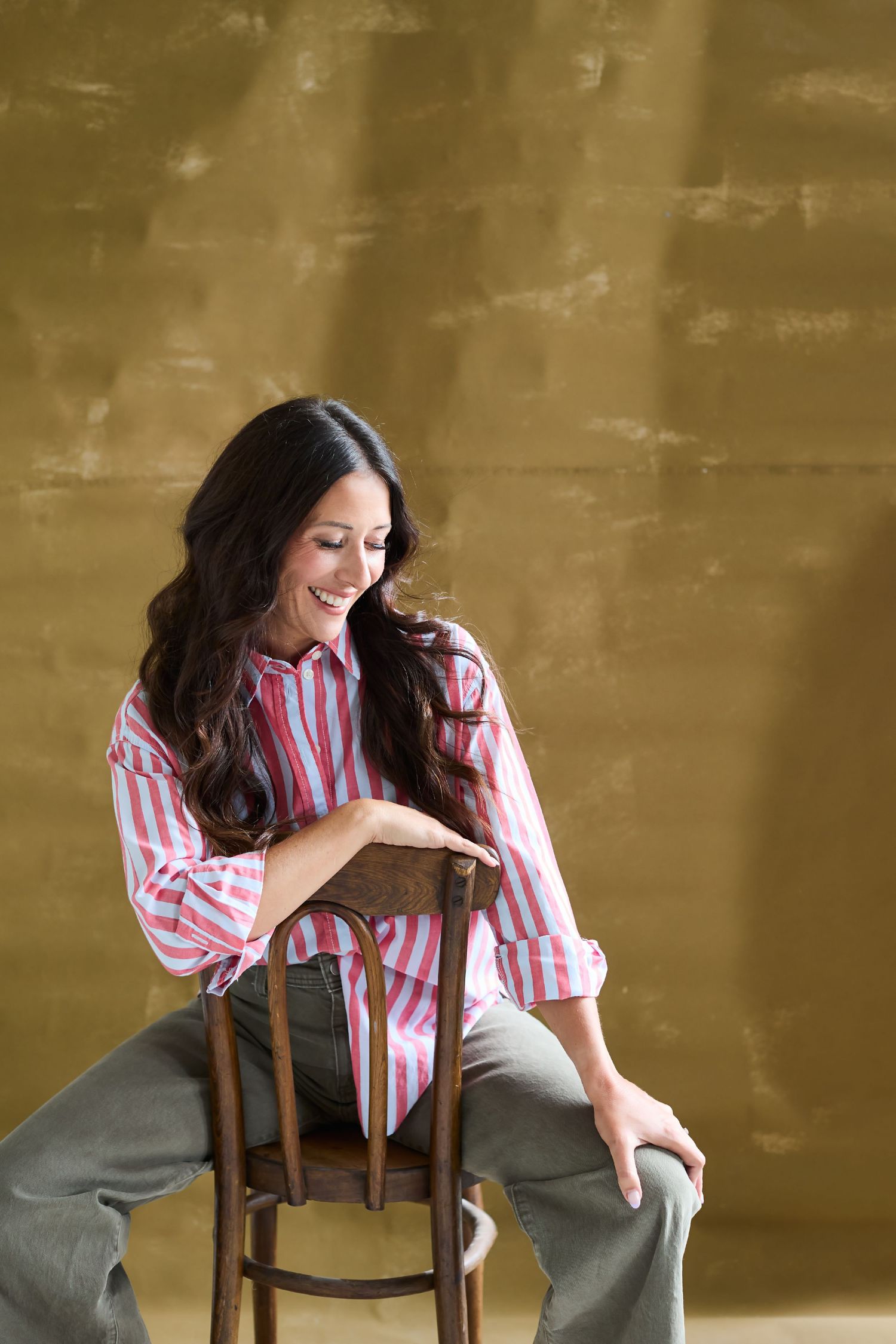 Woman in pink striped shirt sitting on wooden chair, smiling downward against golden brown background.