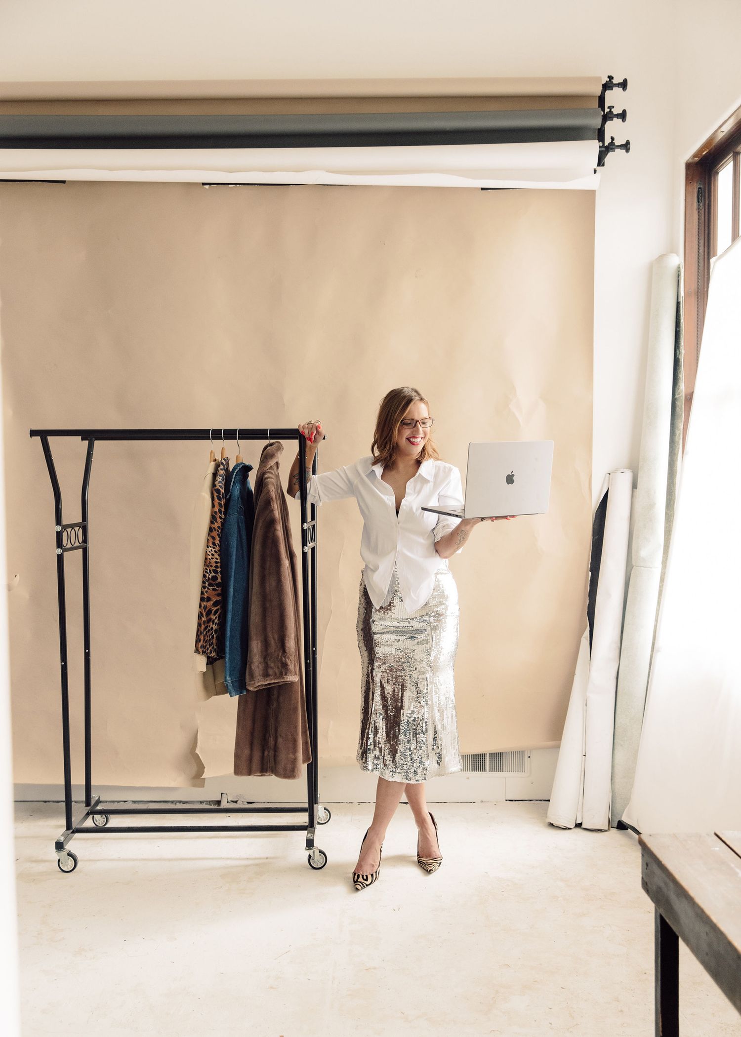 Person in business attire stands next to clothing rack while working on laptop.