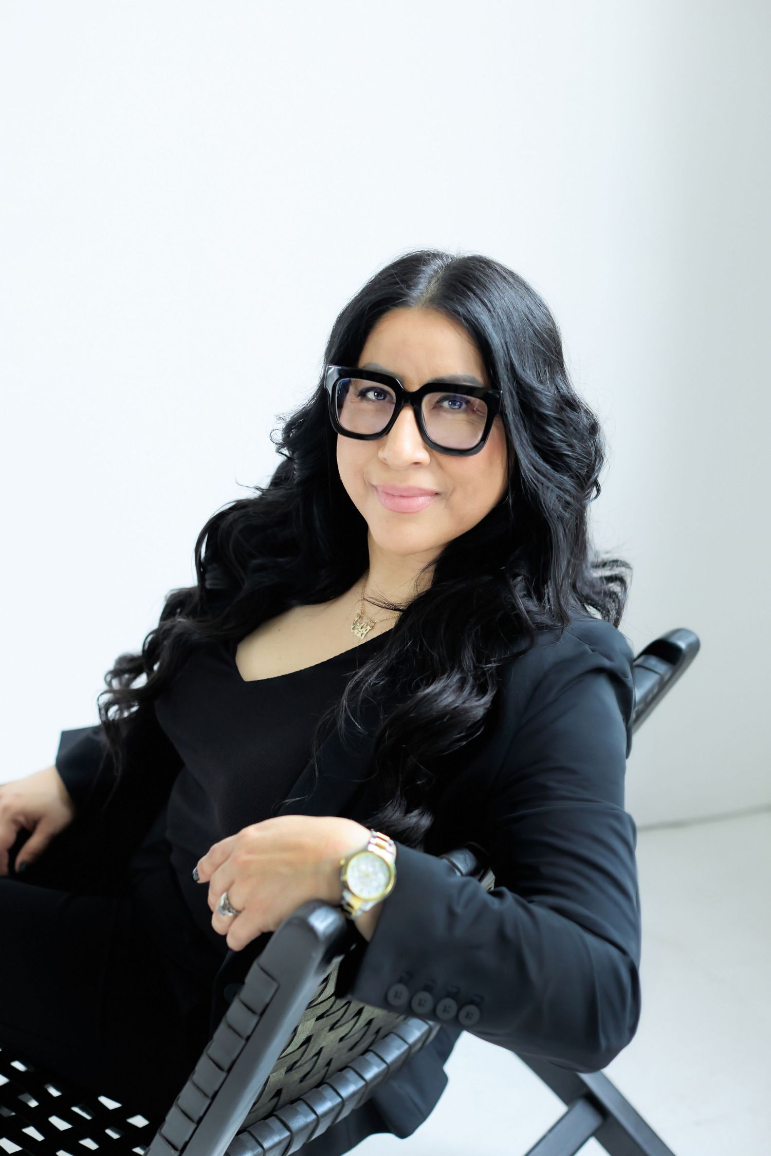 Woman with long black hair and bold glasses in black blazer sitting confidently in chair against white background.