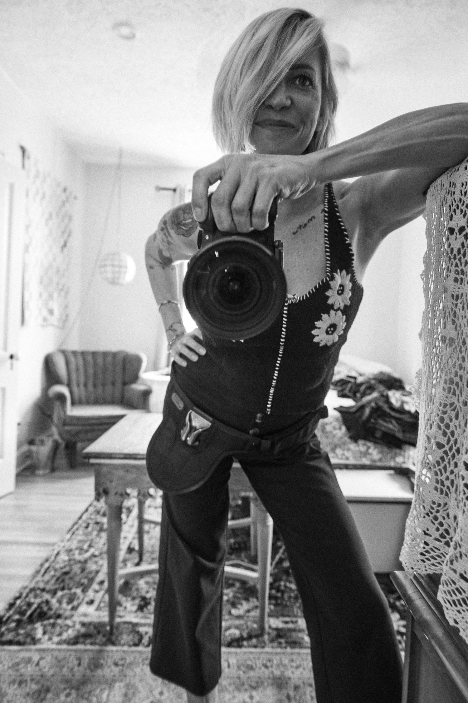 Black and white photo of a tattooed woman taking a mirror selfie with a DSLR camera in a cozy home interior.