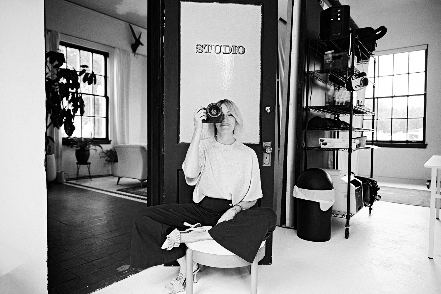 Black and white photo of a blonde woman sitting in a photography studio, holding a camera up to her face.