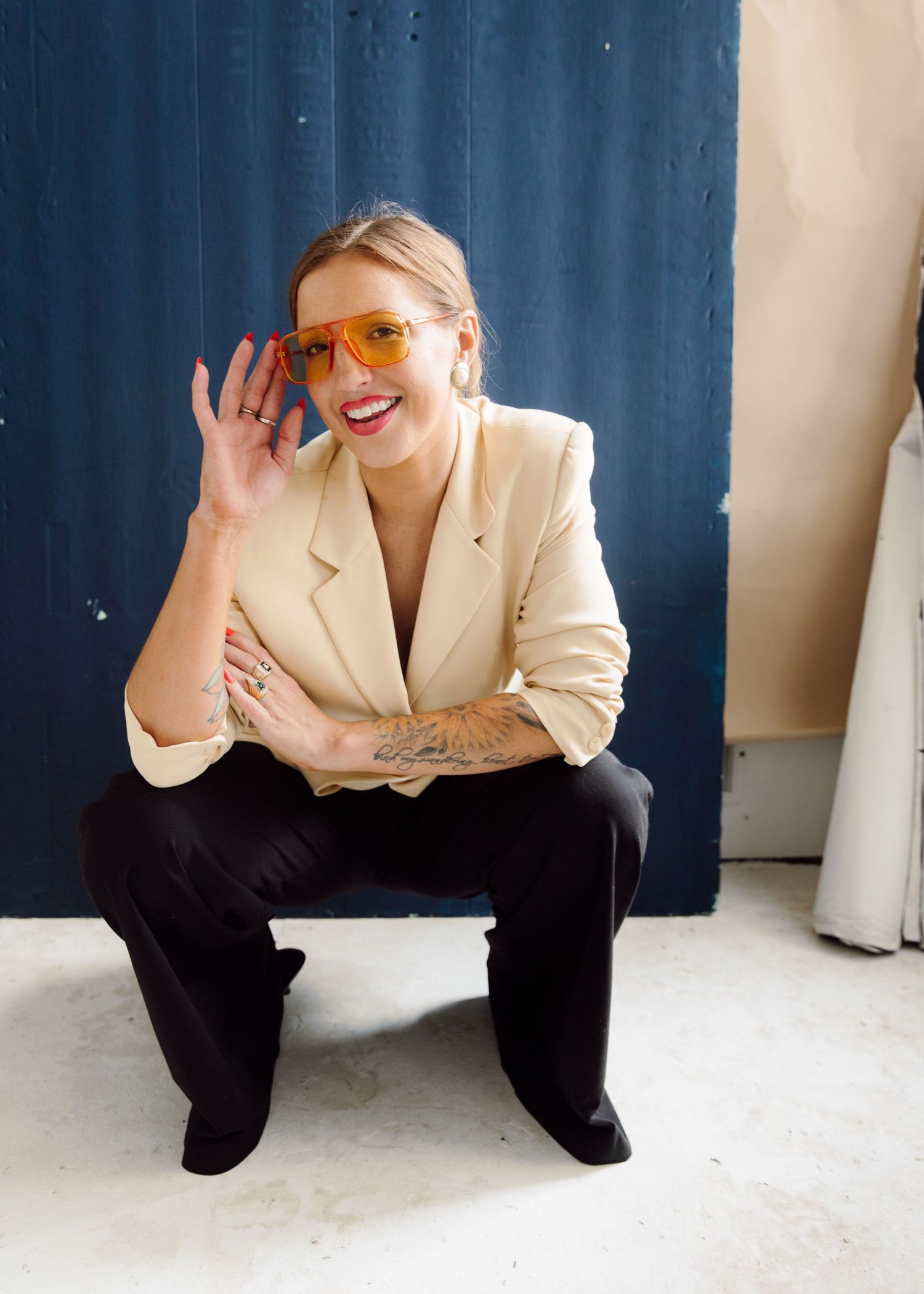 Smiling blonde woman in beige blazer and orange sunglasses crouching playfully, showing tattoos on her arm.