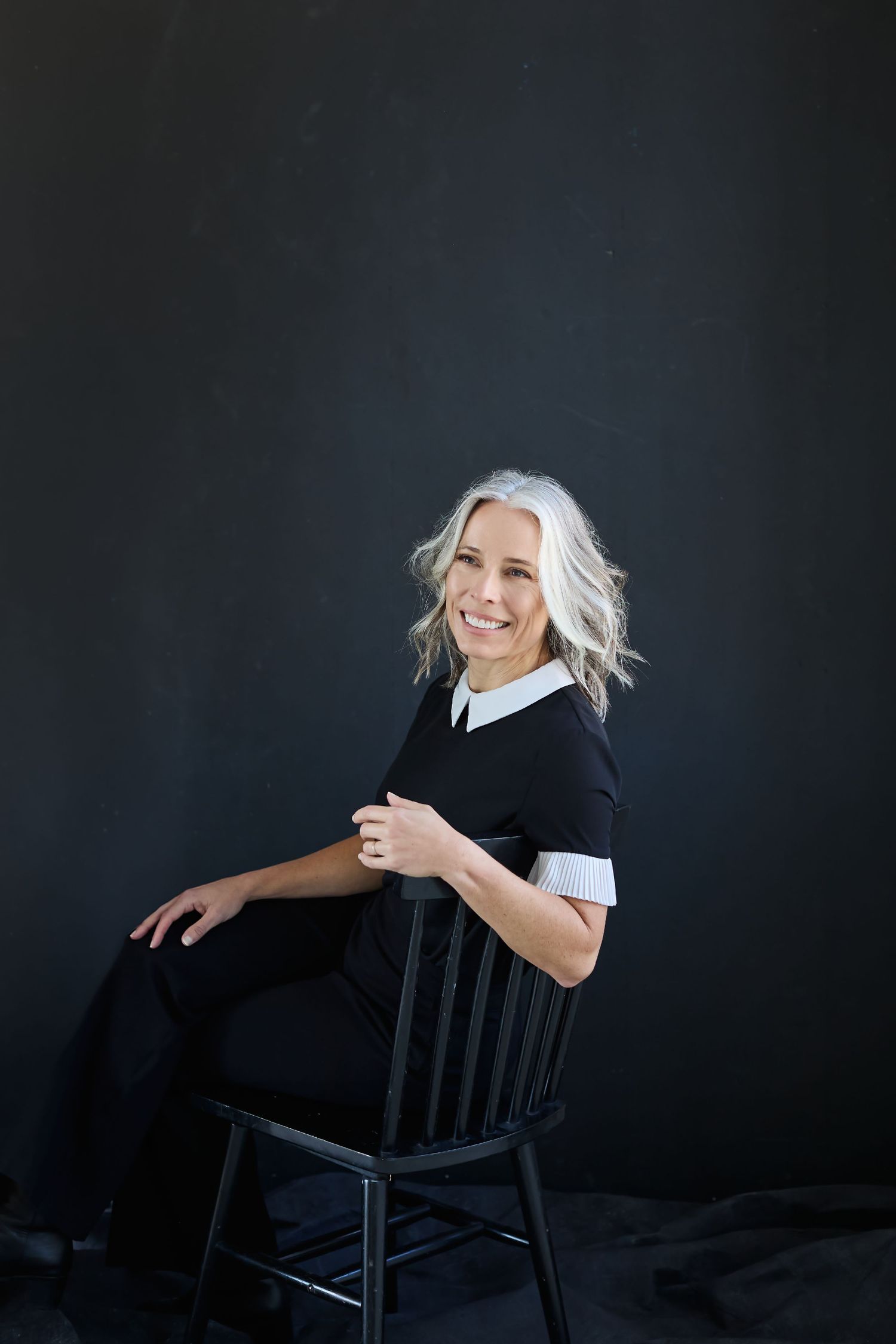 Smiling woman with silver hair sits on black chair, wearing black dress with white collar, against dark background.