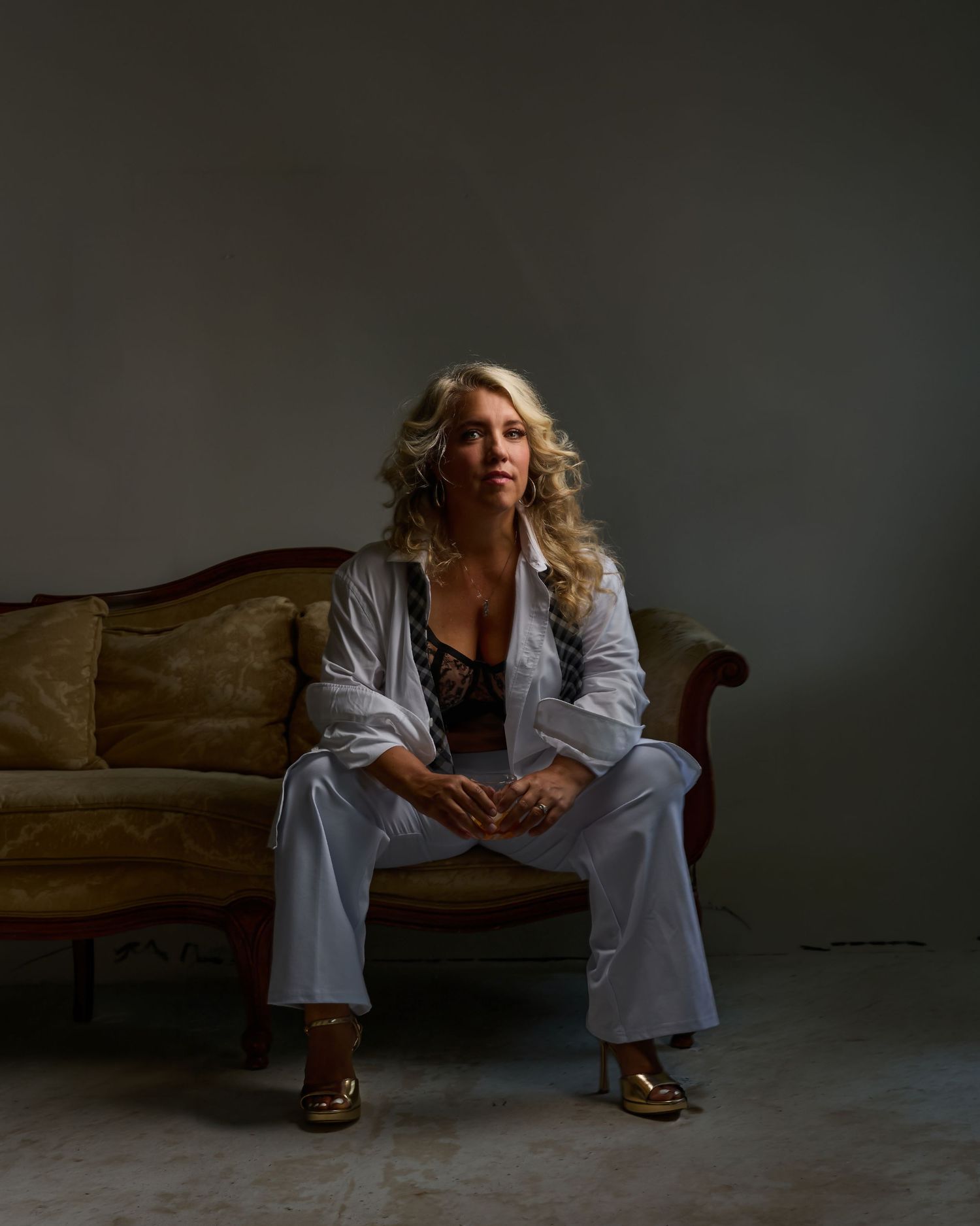 Blonde woman in white pantsuit sits on vintage gold sofa in moody studio portrait setting.