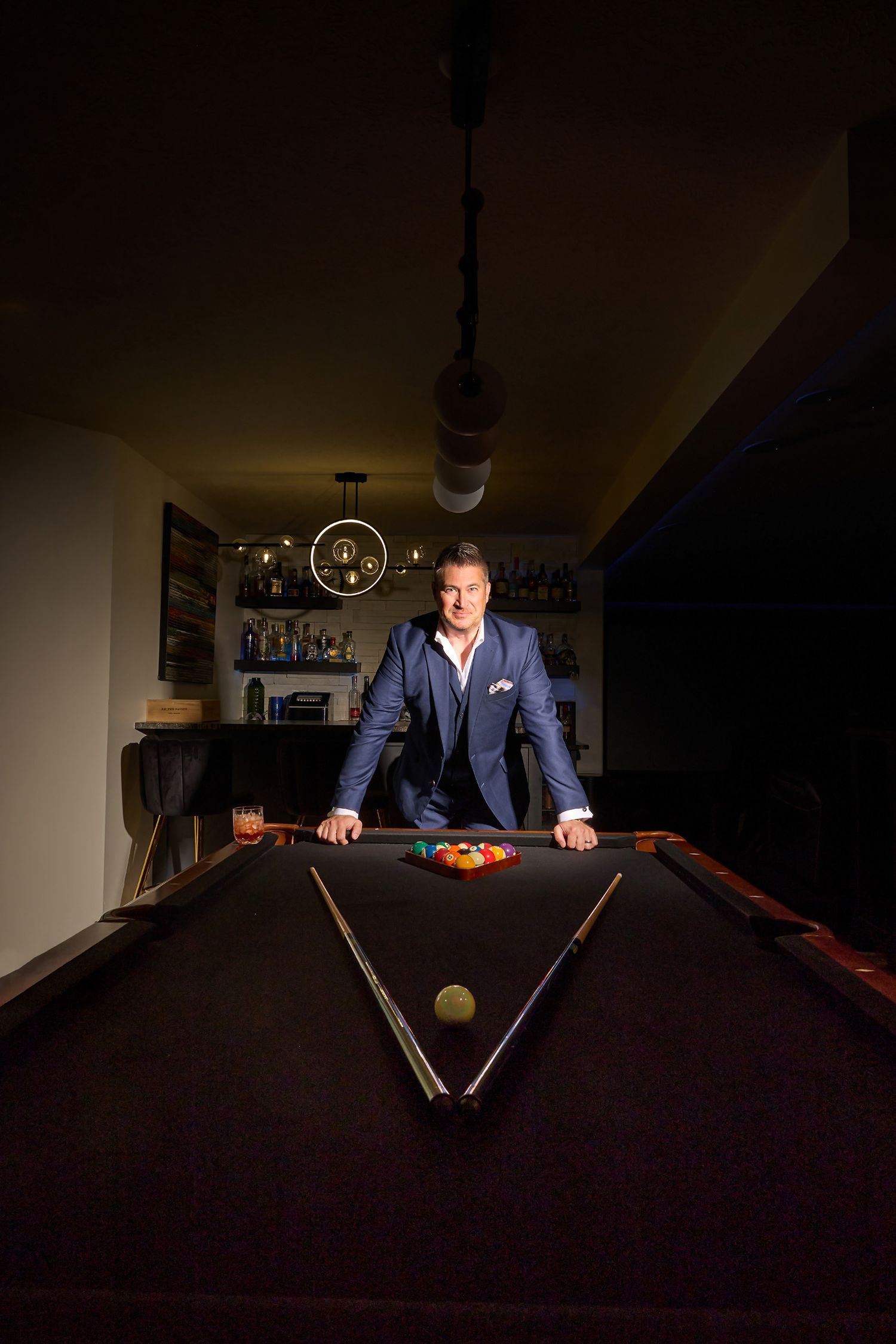 Dramatic lighting illuminates a person playing pool in a dark room wearing a blue suit.