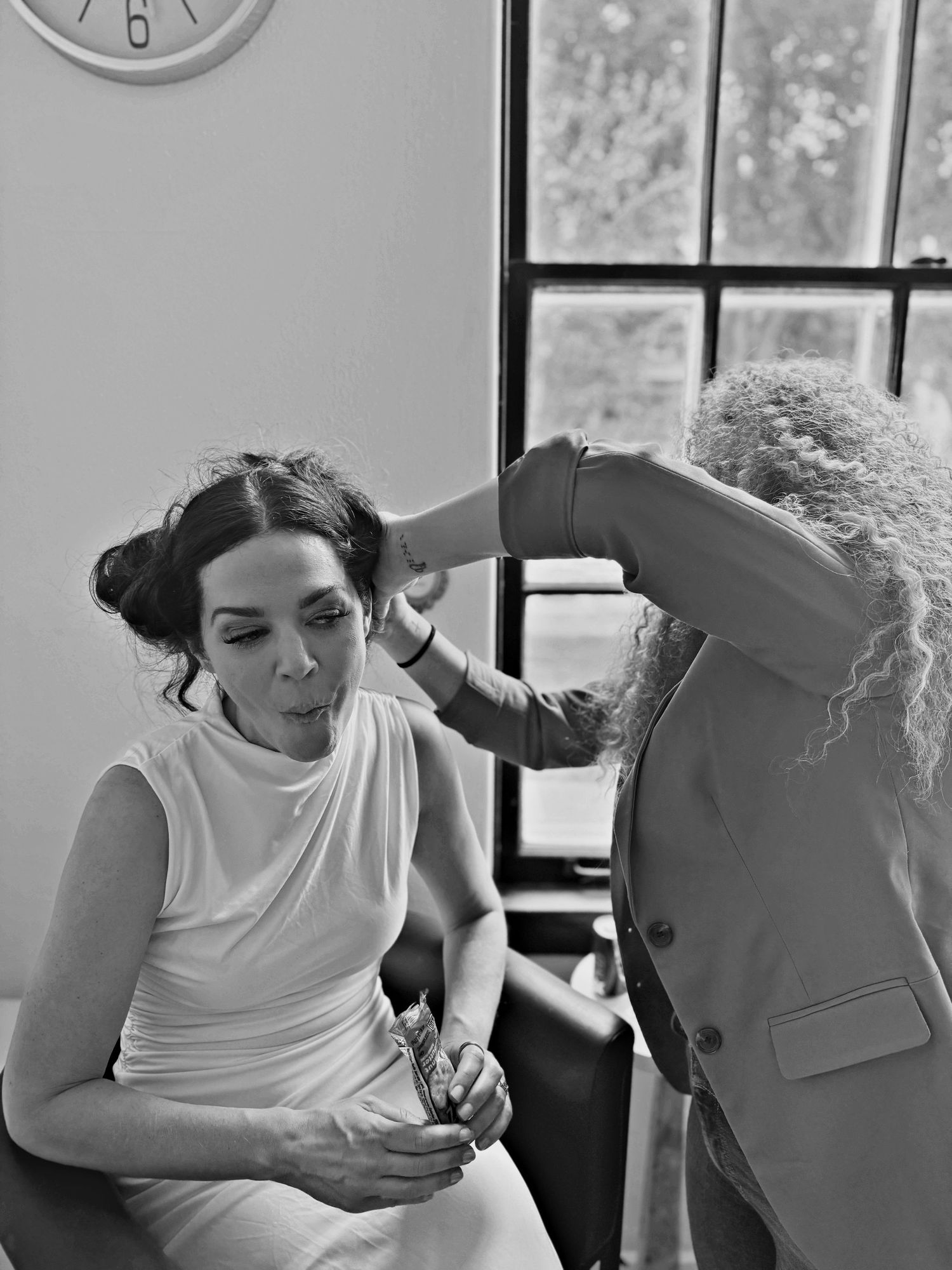 Black and white photo of a woman in white dress getting hair styled by curly-haired stylist near a window.