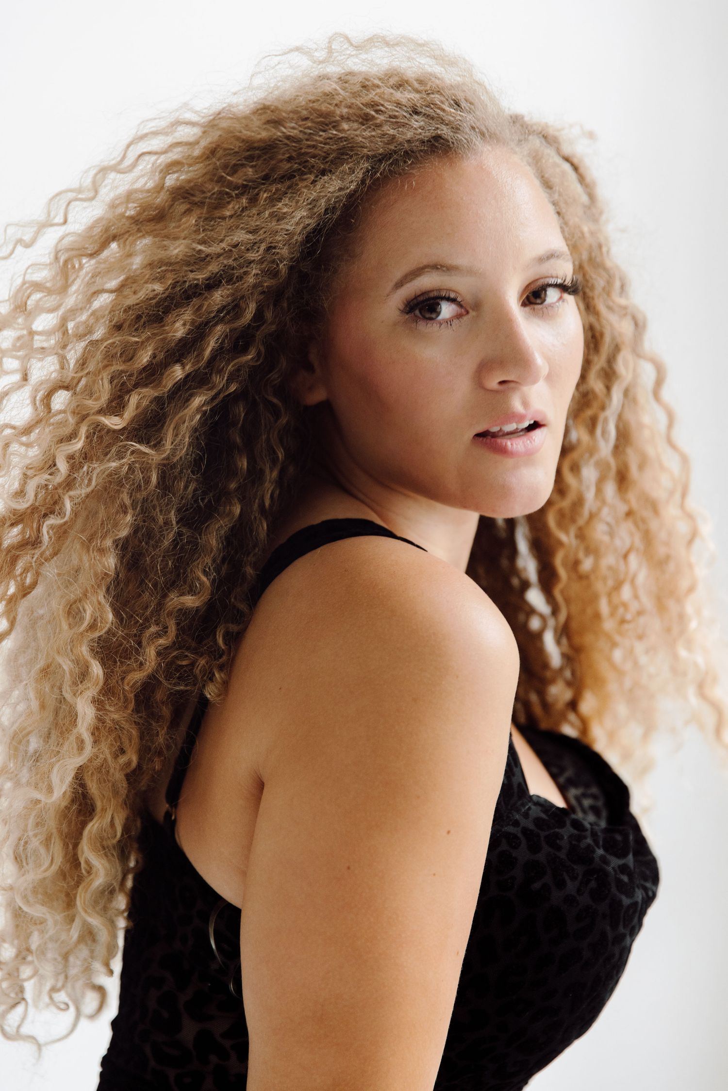 Young woman with voluminous curly blonde hair wearing a black top, posing against a white background.