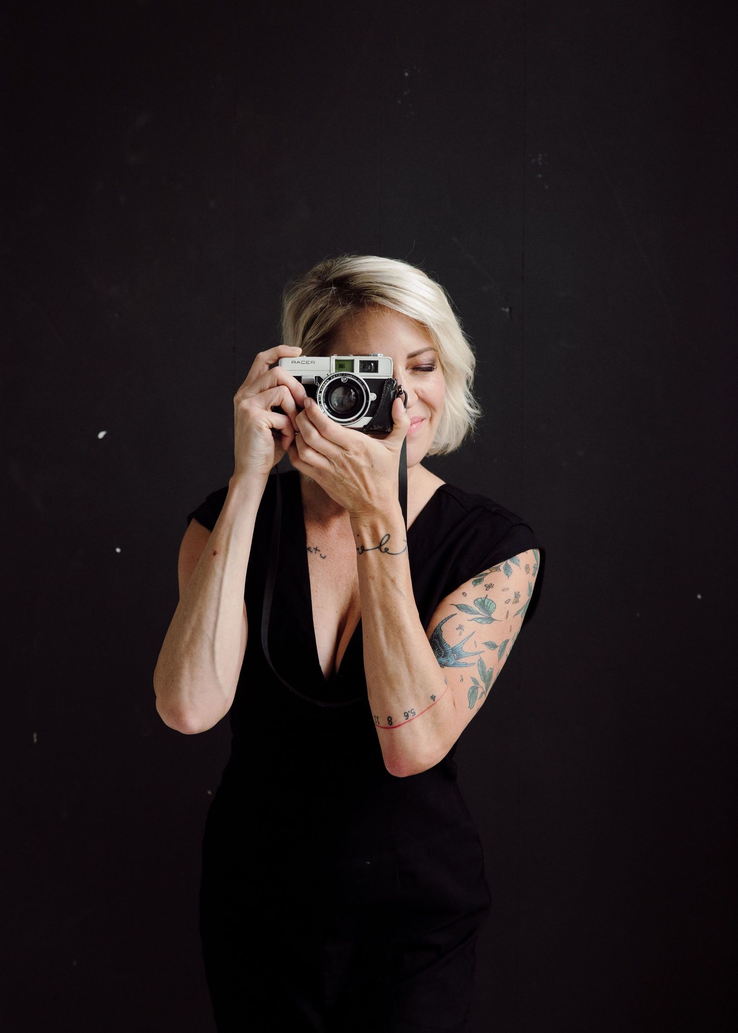 Blonde woman with tattoos in black dress holds a film camera up to her face, smiling, against dark background.