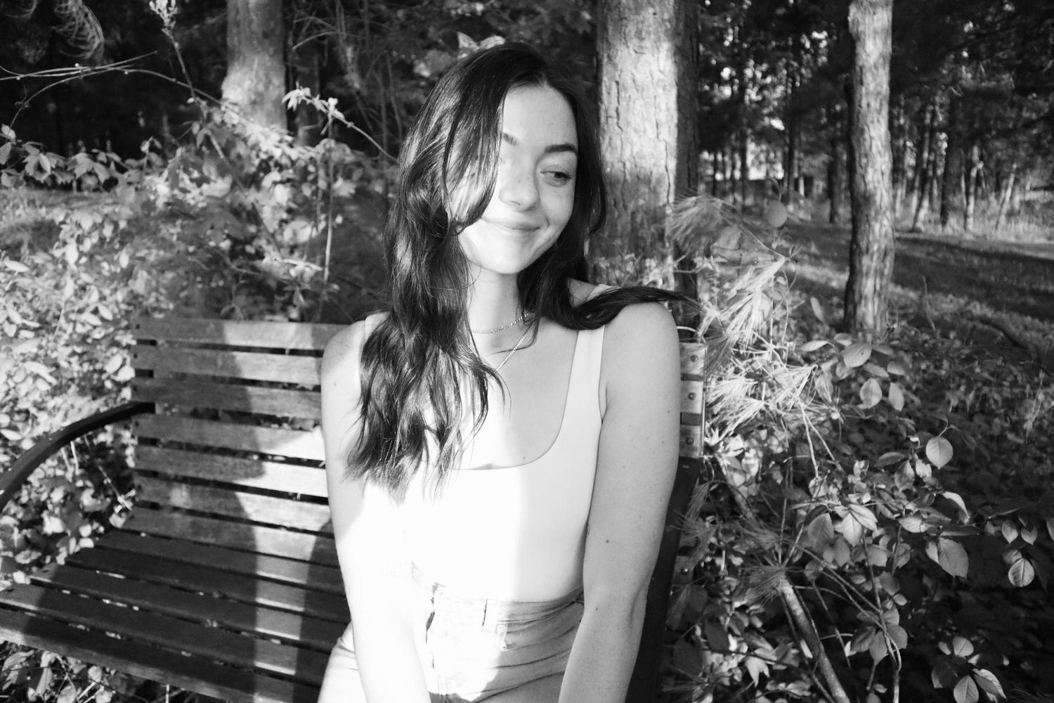 Black and white portrait of a woman in a white sleeveless top sitting on a park bench surrounded by foliage.