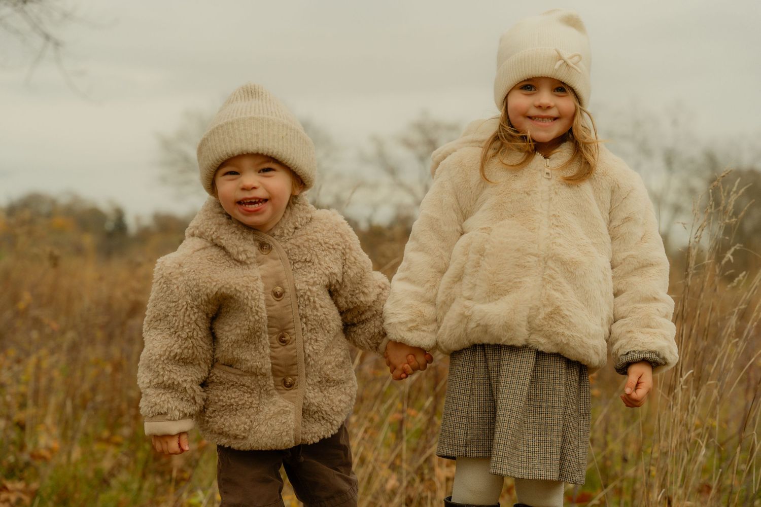 Two children wearing cozy beige winter coats and matching knit beanies stand together in a golden autumn field.