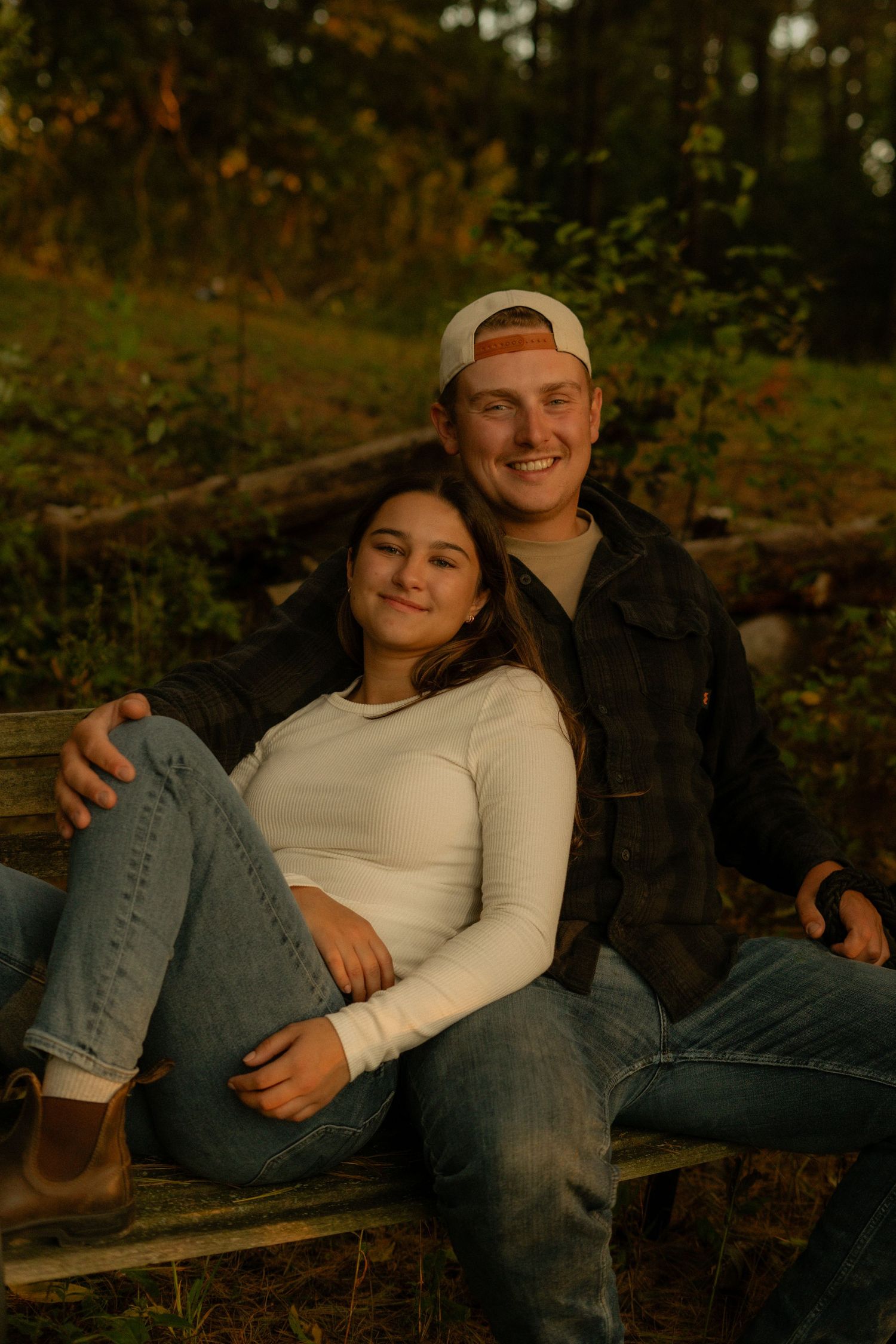 A couple sits together on a log at sunset in a wooded area, wearing casual clothing and sharing an intimate moment.