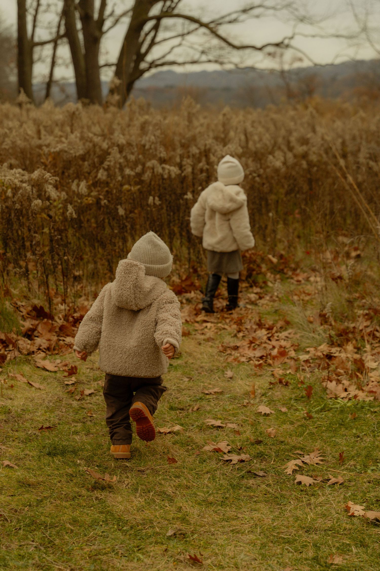 Two bundled-up figures in cozy beige coats explore an autumn meadow with tall dried grasses and mountains in the distance.