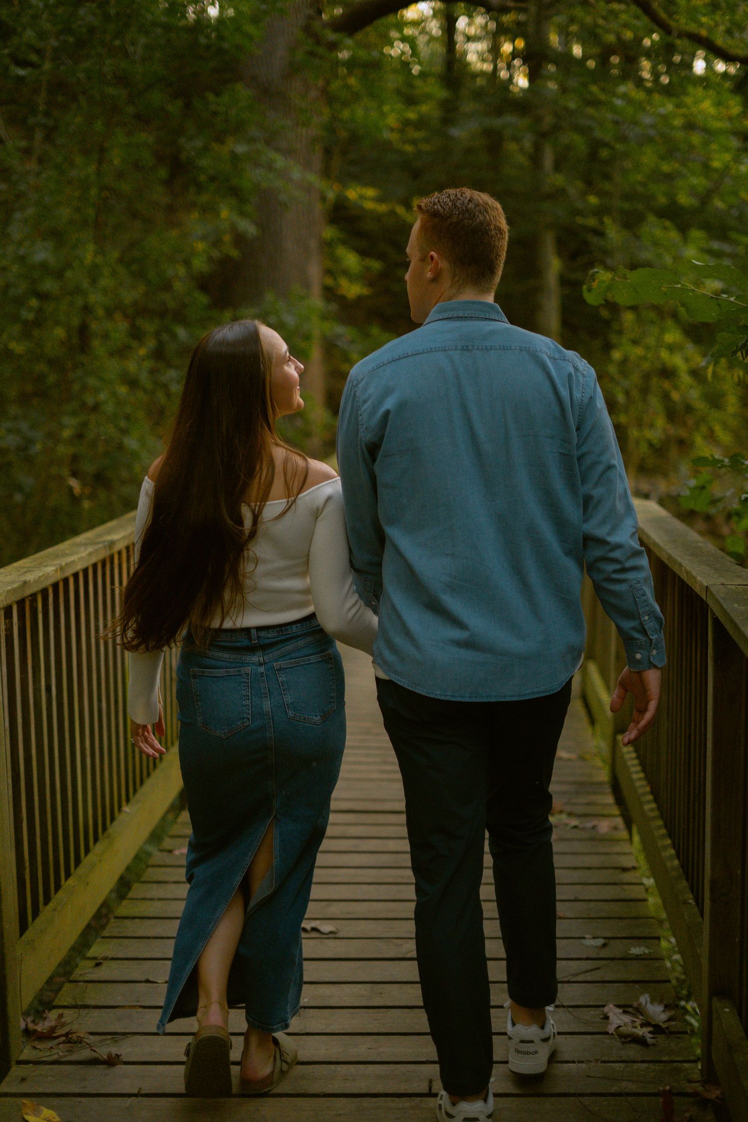 A couple walking together on a wooden footbridge in a lush forest during golden hour lighting.